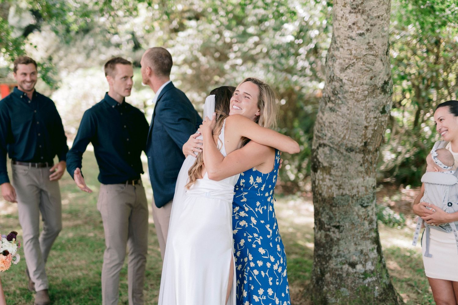 People mingling at an outdoor wedding reception beneath large trees.