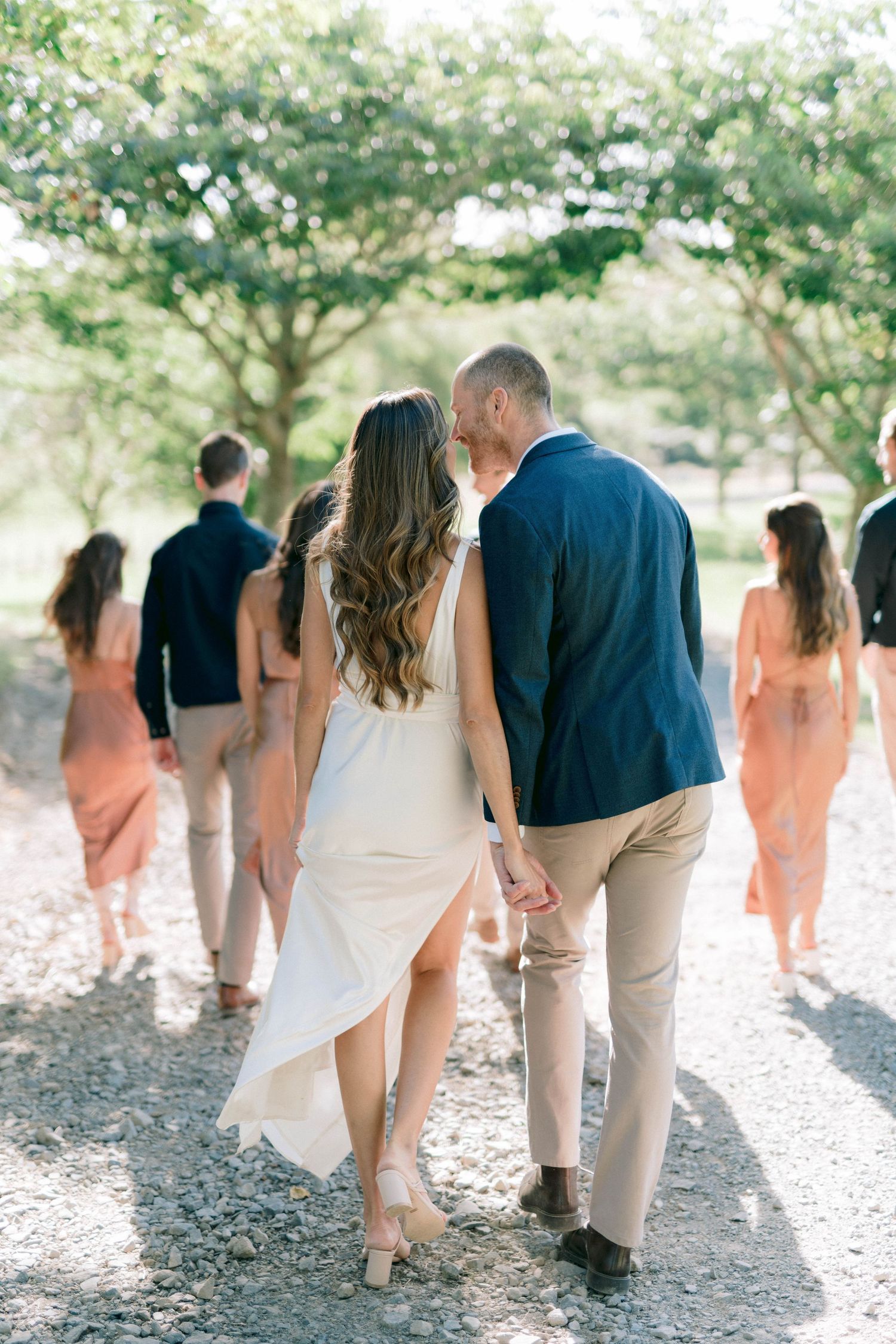 Wedding party wearing coral dresses and khaki suits walk along a tree-lined path during outdoor photo session.