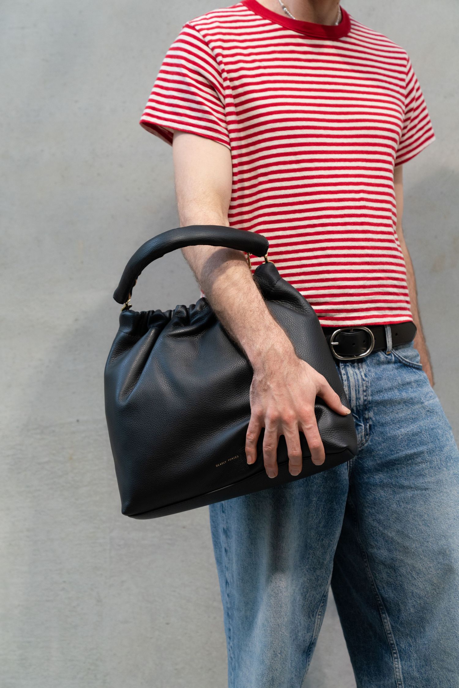 Casual outfit with red and white striped t-shirt, blue jeans and black leather gathered handbag against gray wall.