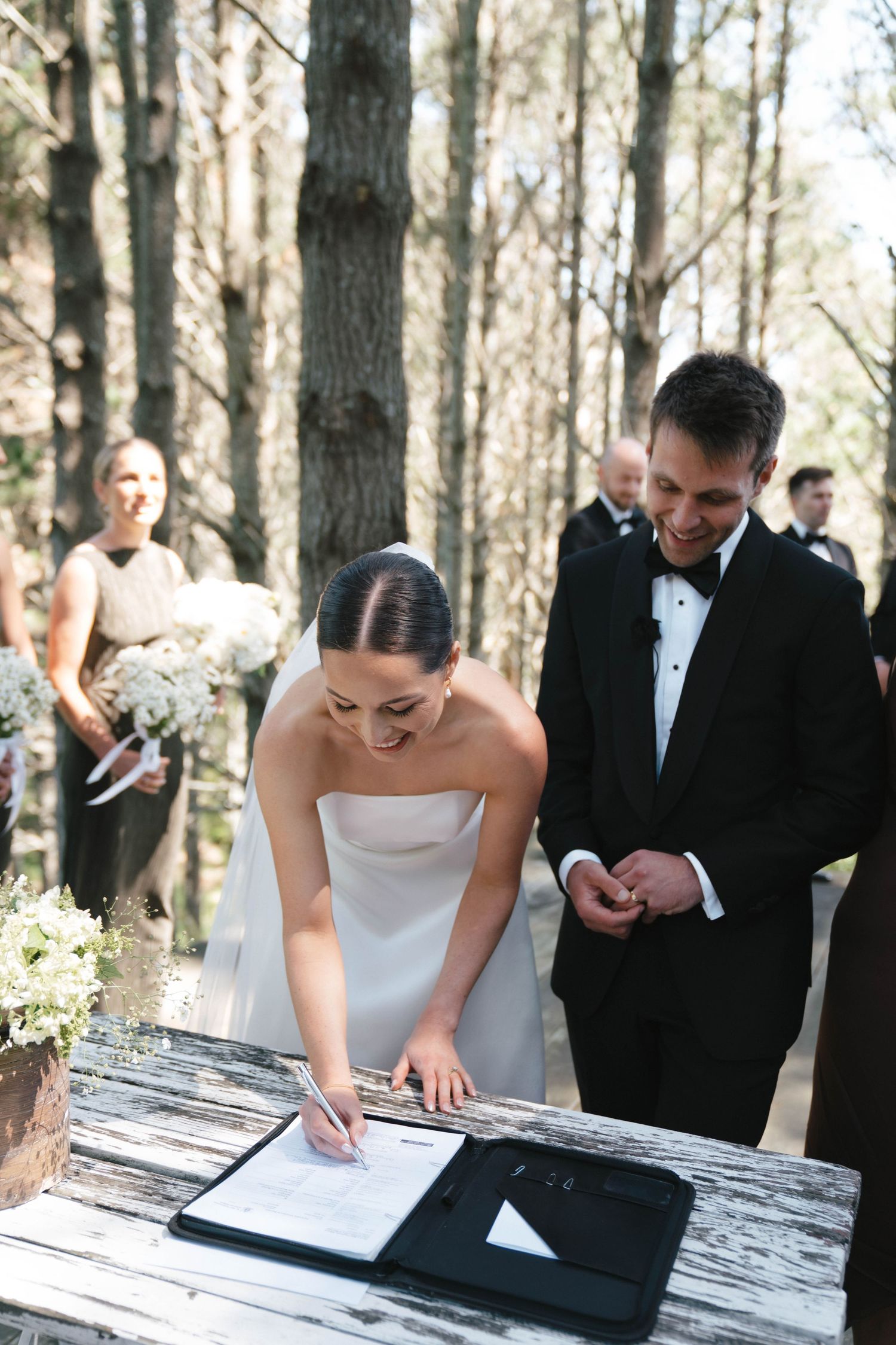 A couple in formal wedding attire signing documents on a rustic wooden table in a forest setting with guests looking on.