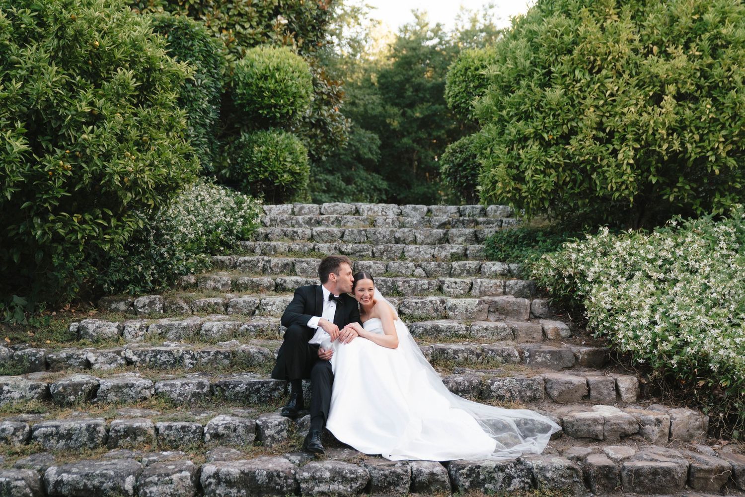 A couple in wedding attire sits together on rustic stone steps surrounded by lush greenery in an outdoor garden setting.