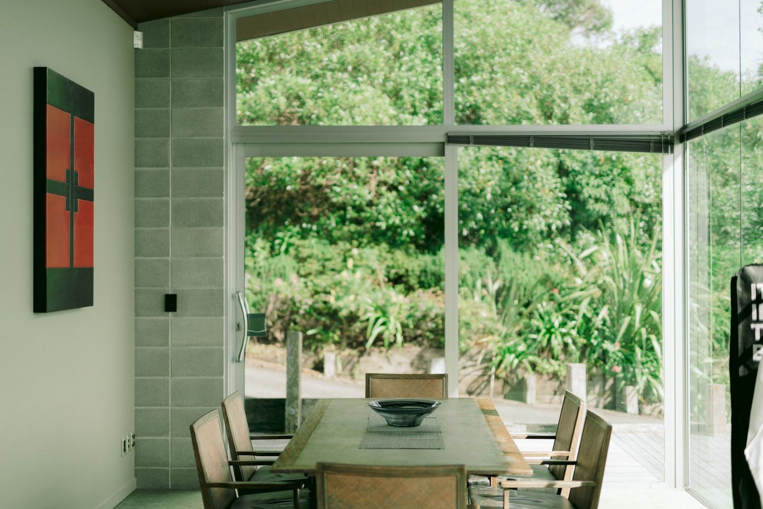 Modern dining area with wooden table and chairs against large windows overlooking lush green garden.