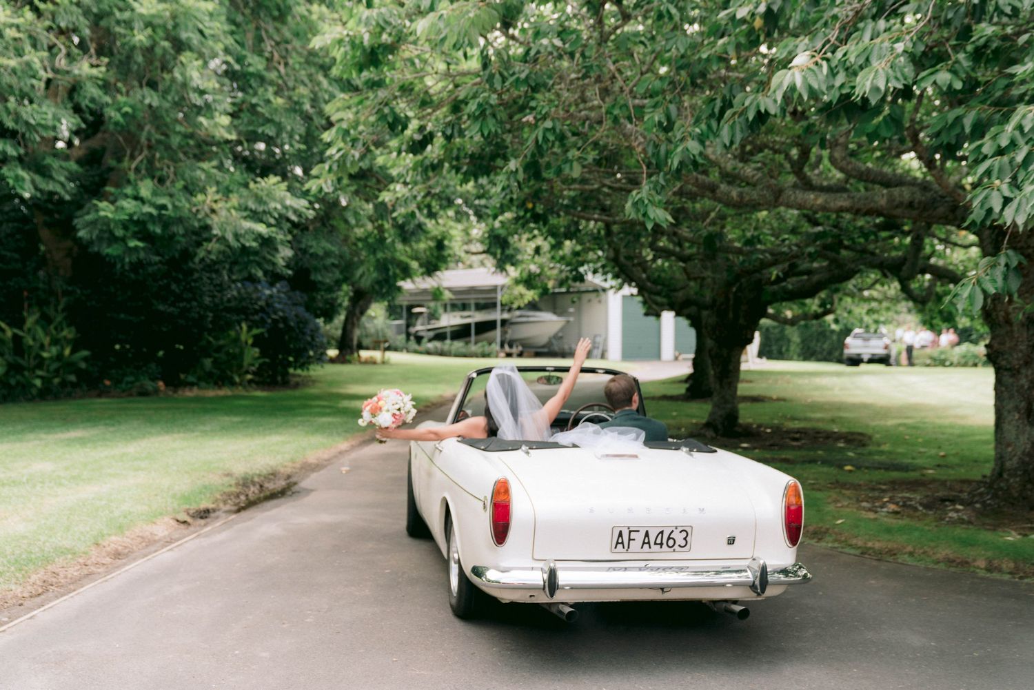 A classic convertible car drives down a tree-lined residential street on a sunny day.