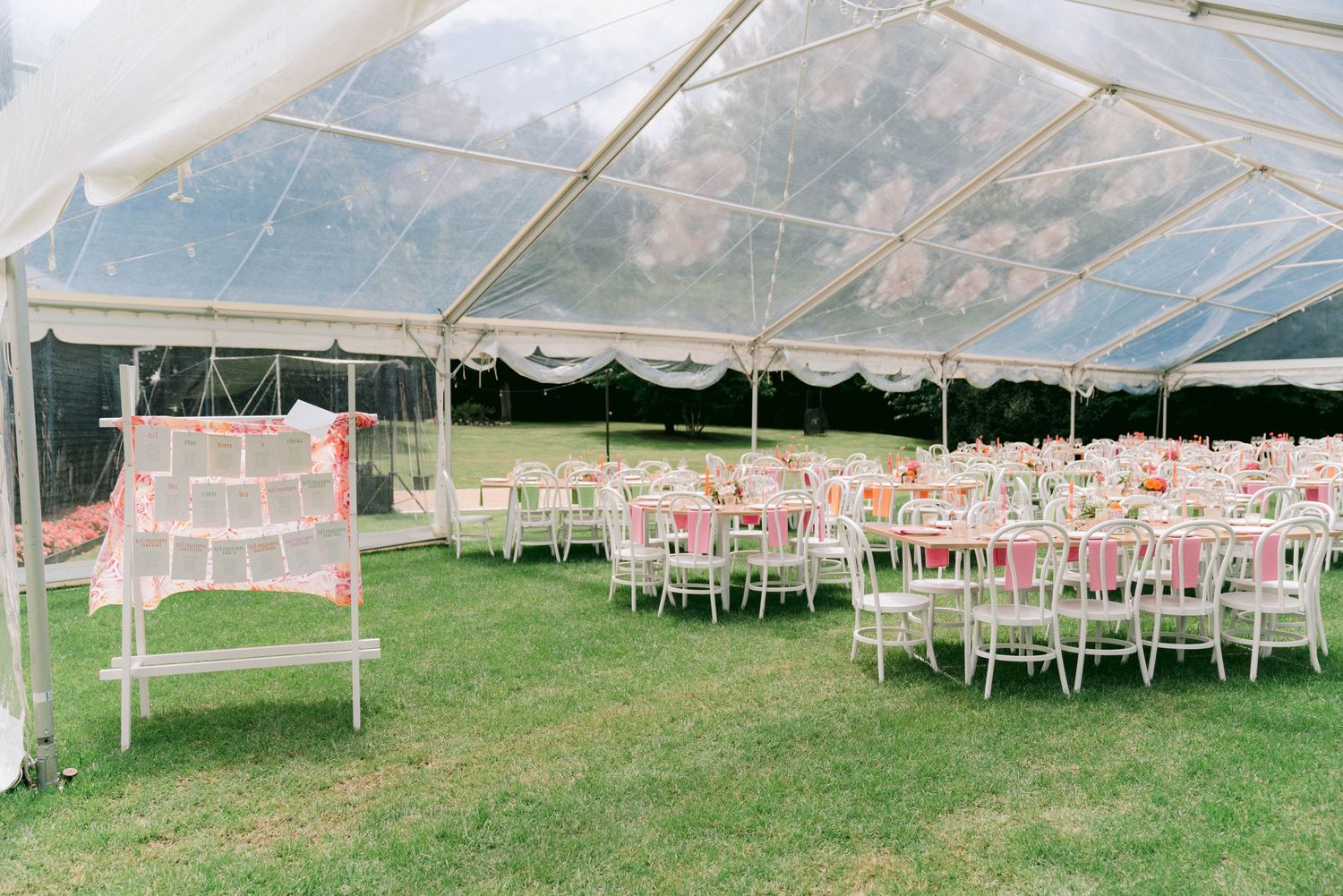 Wedding reception setup under a clear tent with pink and white decor and round tables.