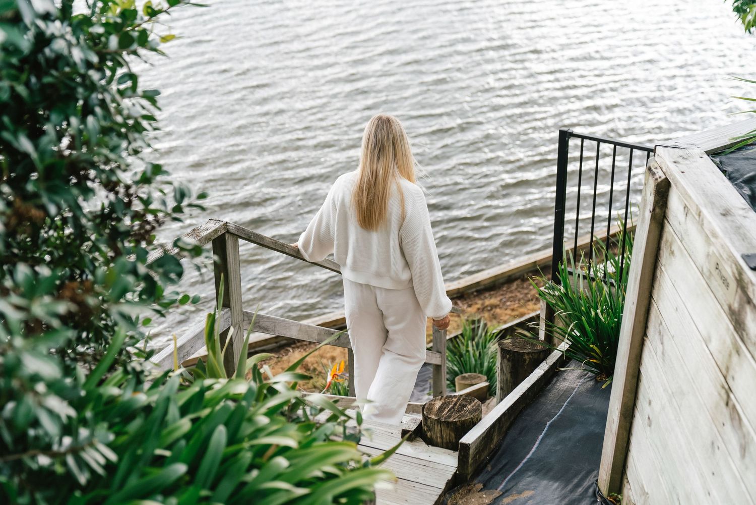 Person in white outfit standing on wooden deck stairs overlooking waterfront.