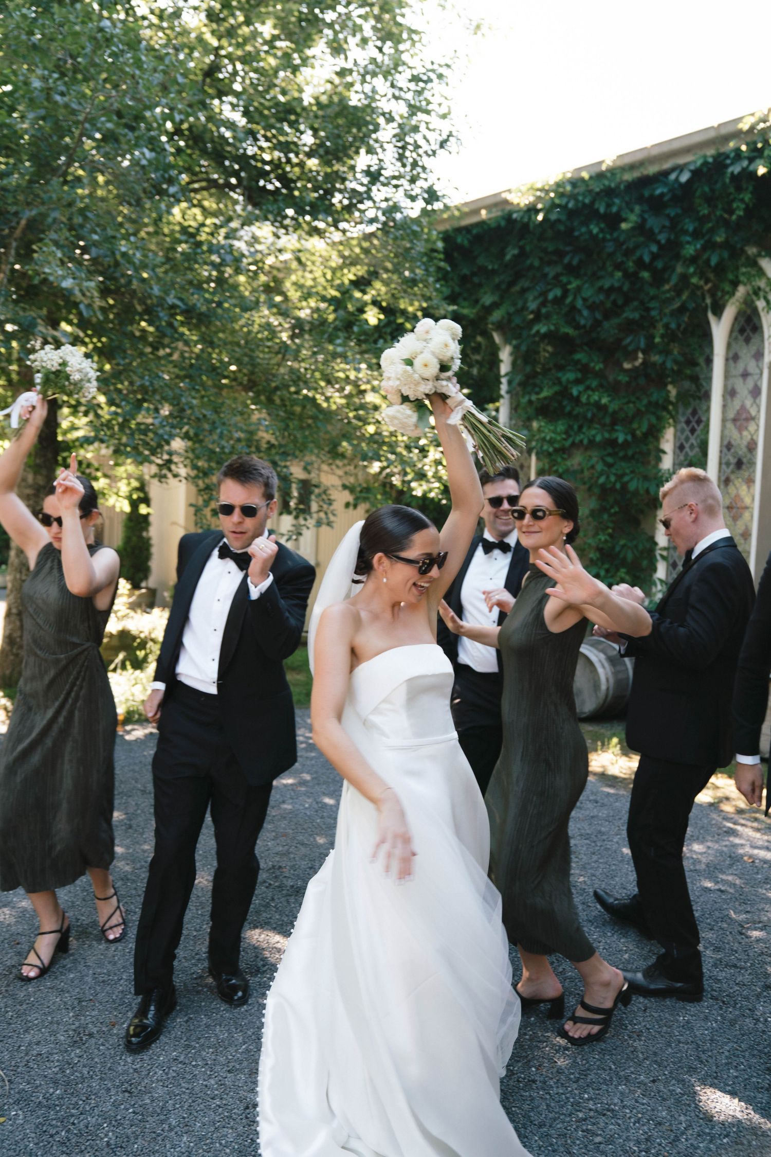 Wedding party in formal attire dances and celebrates on a paved outdoor area surrounded by trees.