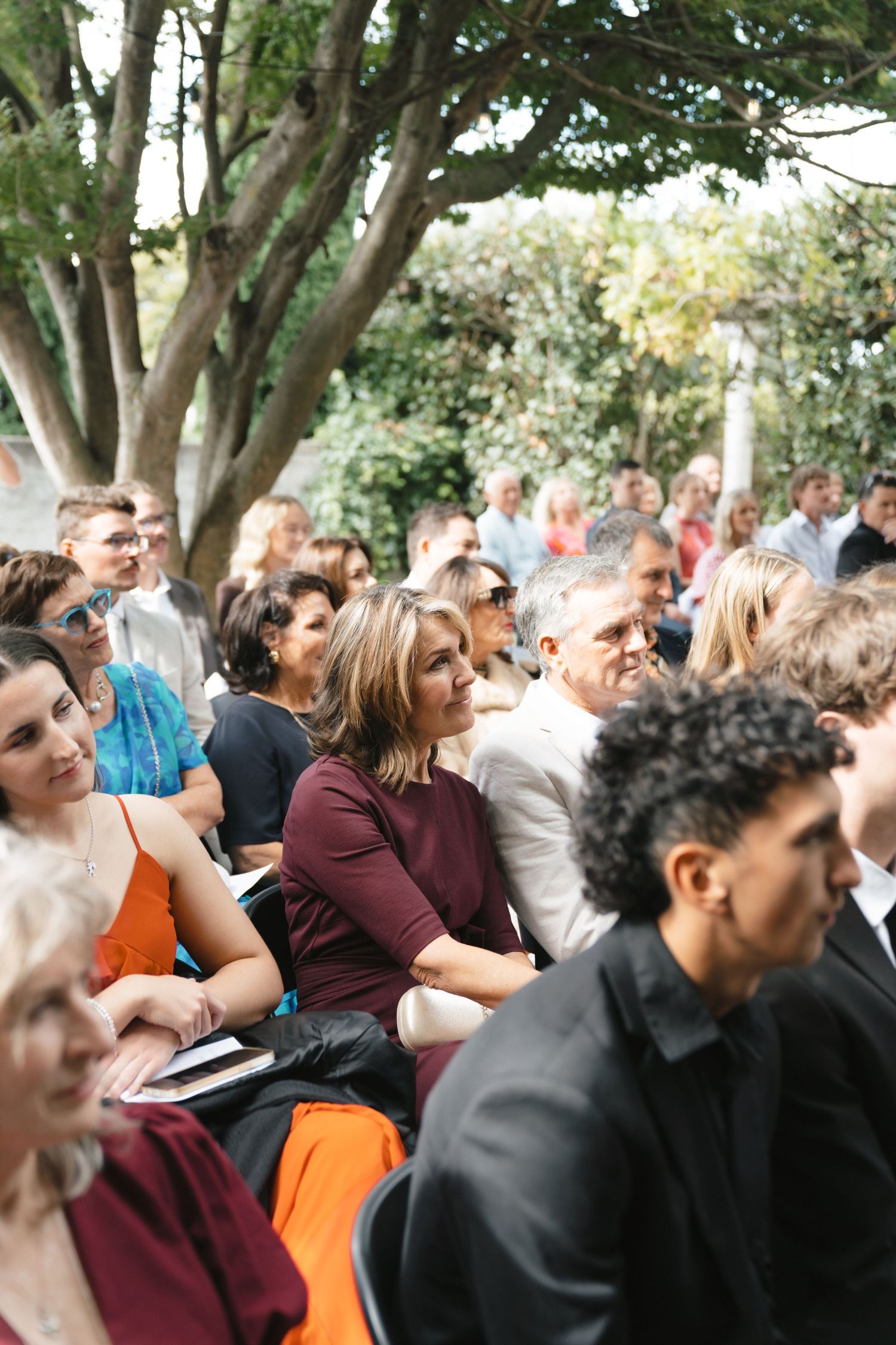Guests seated outdoors at wedding ceremony under large tree canopy.