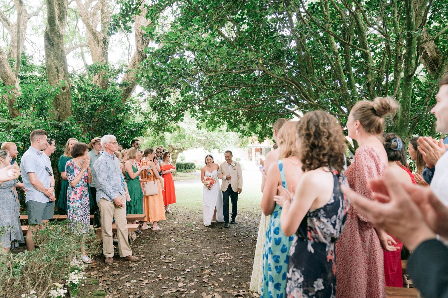 Wedding guests gather for an outdoor ceremony under a canopy of green trees.
