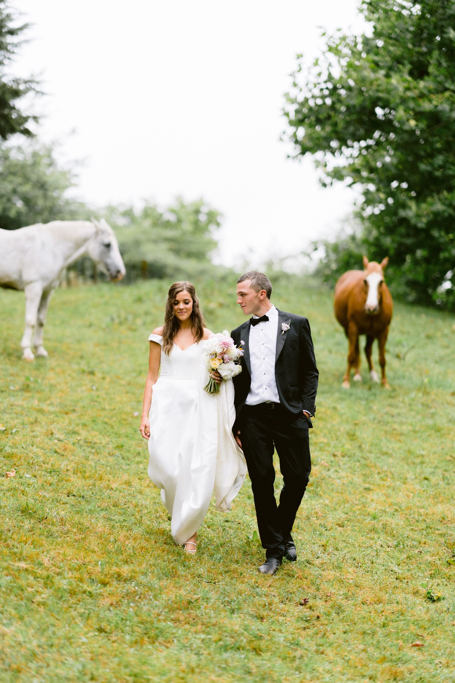 Newlyweds walk through grassy field with horses grazing in the background on their wedding day.