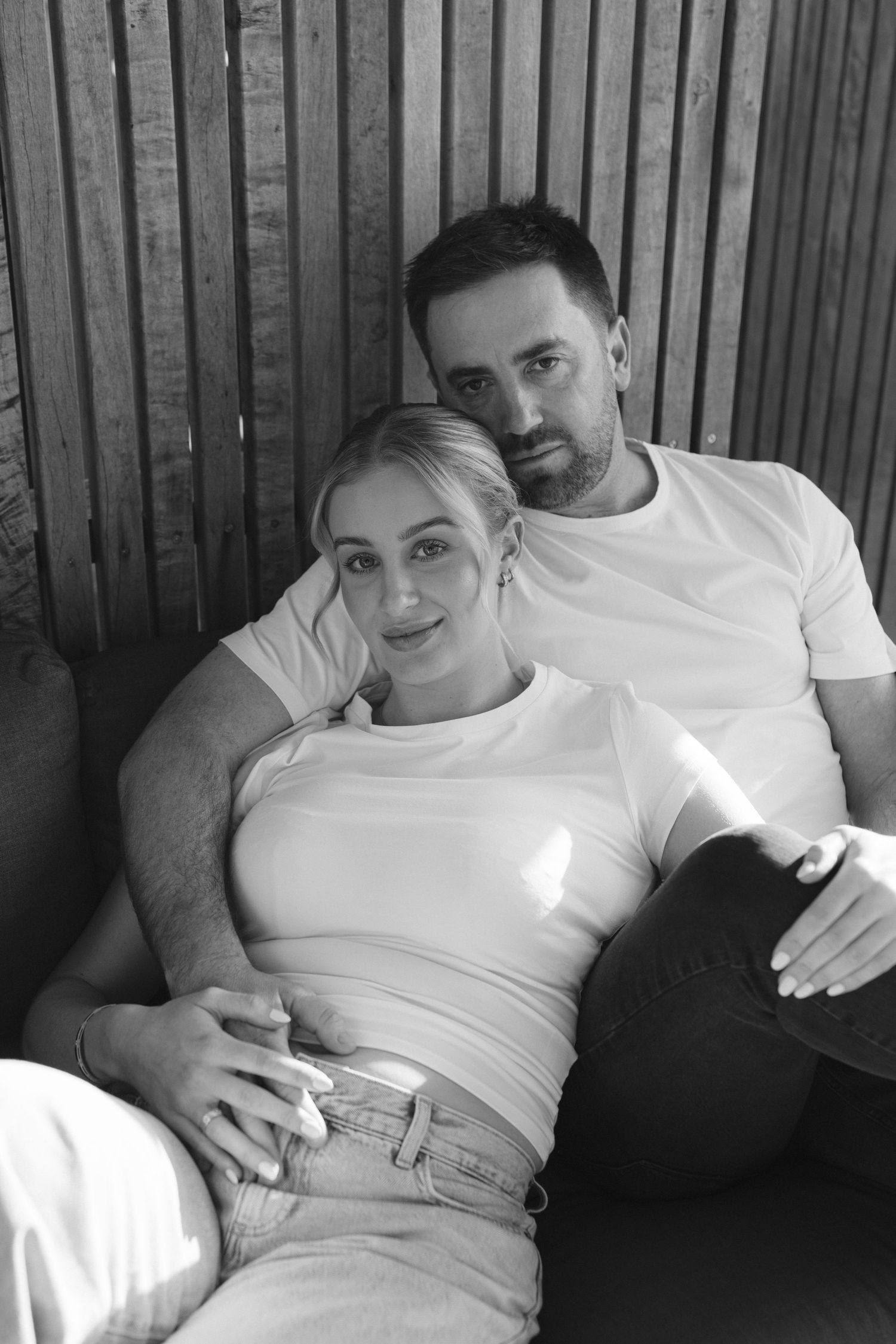 Black and white intimate portrait of a couple relaxing together in casual white tops against a striped wall.