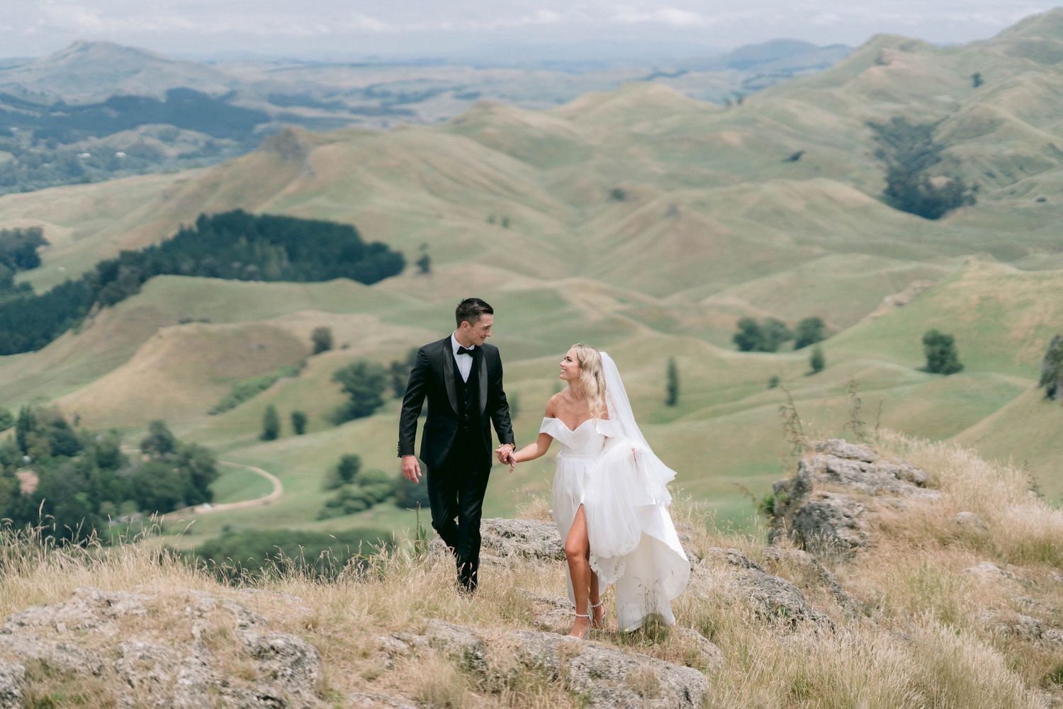 Couple walking hand in hand across a scenic mountain ridge in wedding attire.