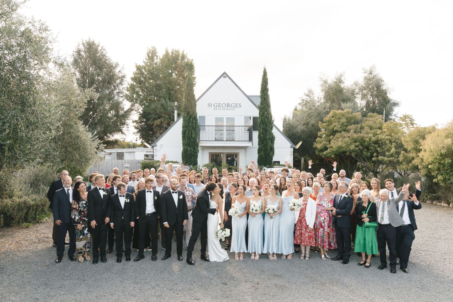Large wedding party group photo taken outdoors with classic white building in background.