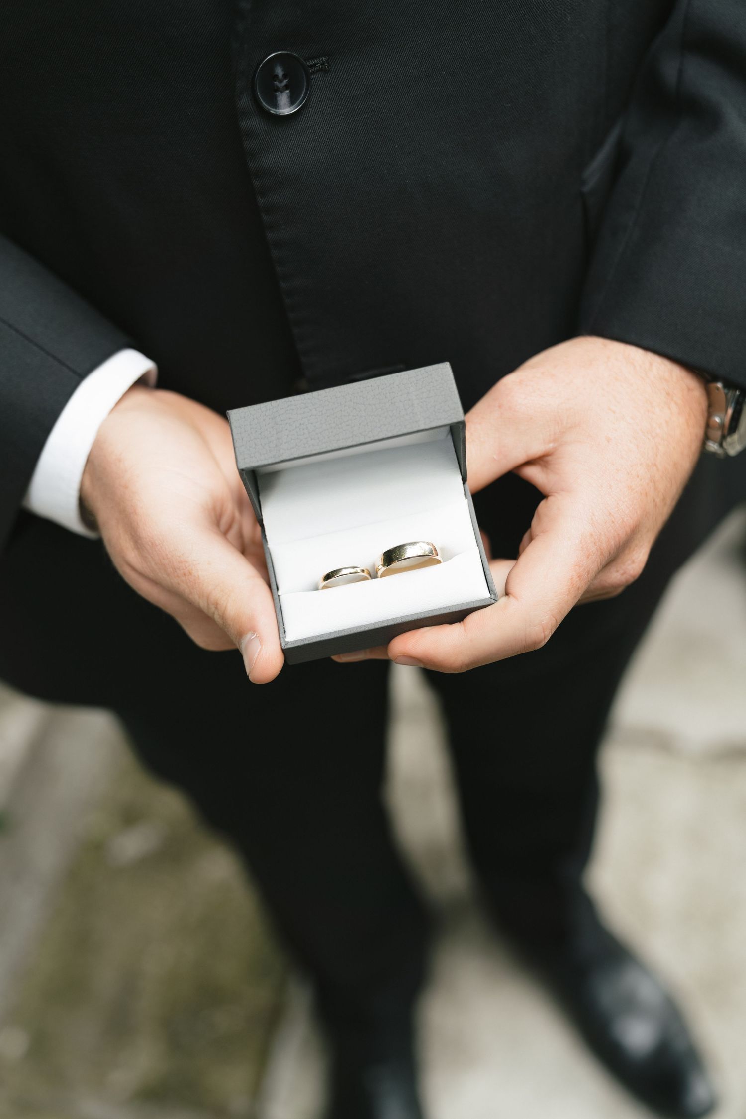 A person in a dark suit holds a small silver box containing a gold wedding ring.