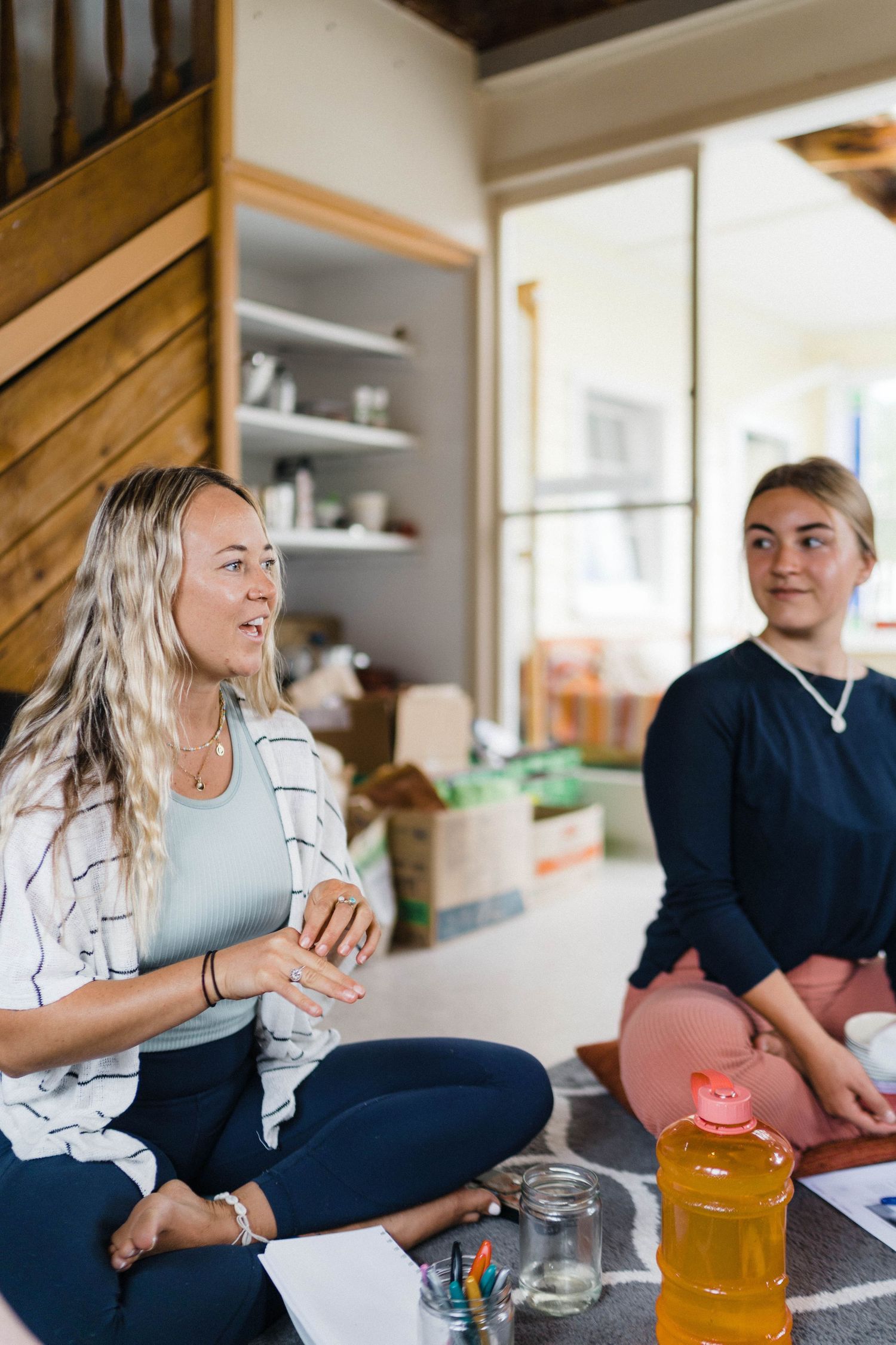 Two people sitting cross-legged on floor during casual indoor gathering with drinks.