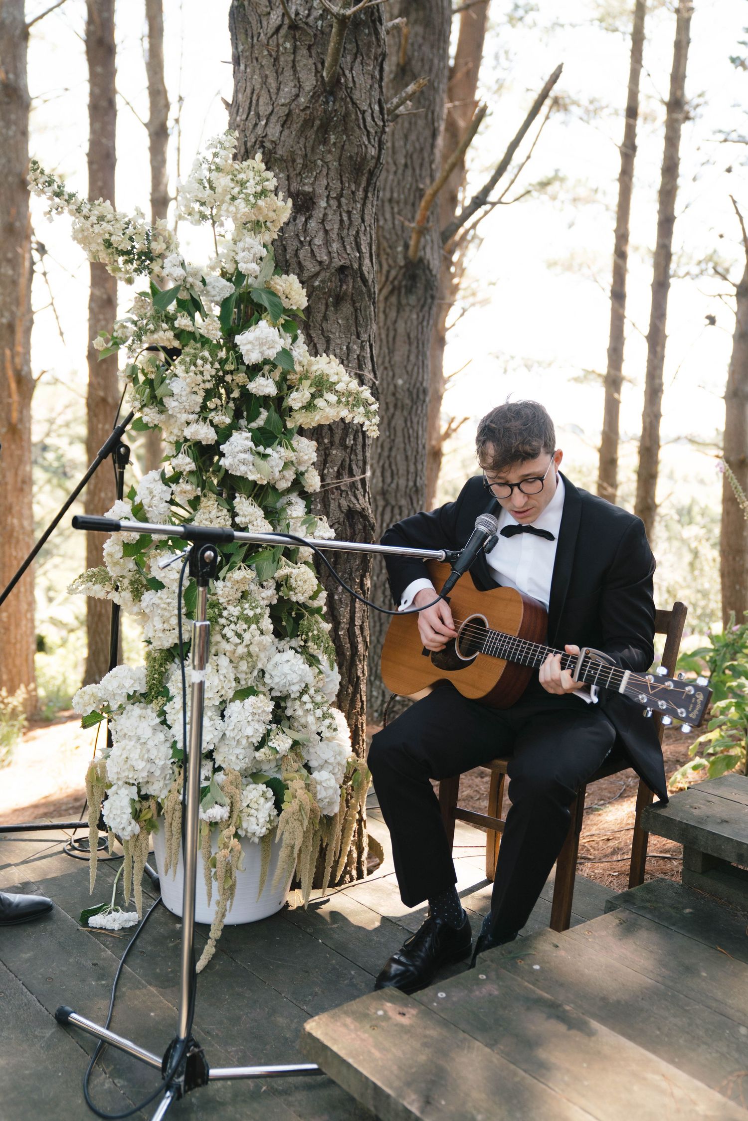 A guitarist in formal attire performs among floral decorations at an outdoor wedding.