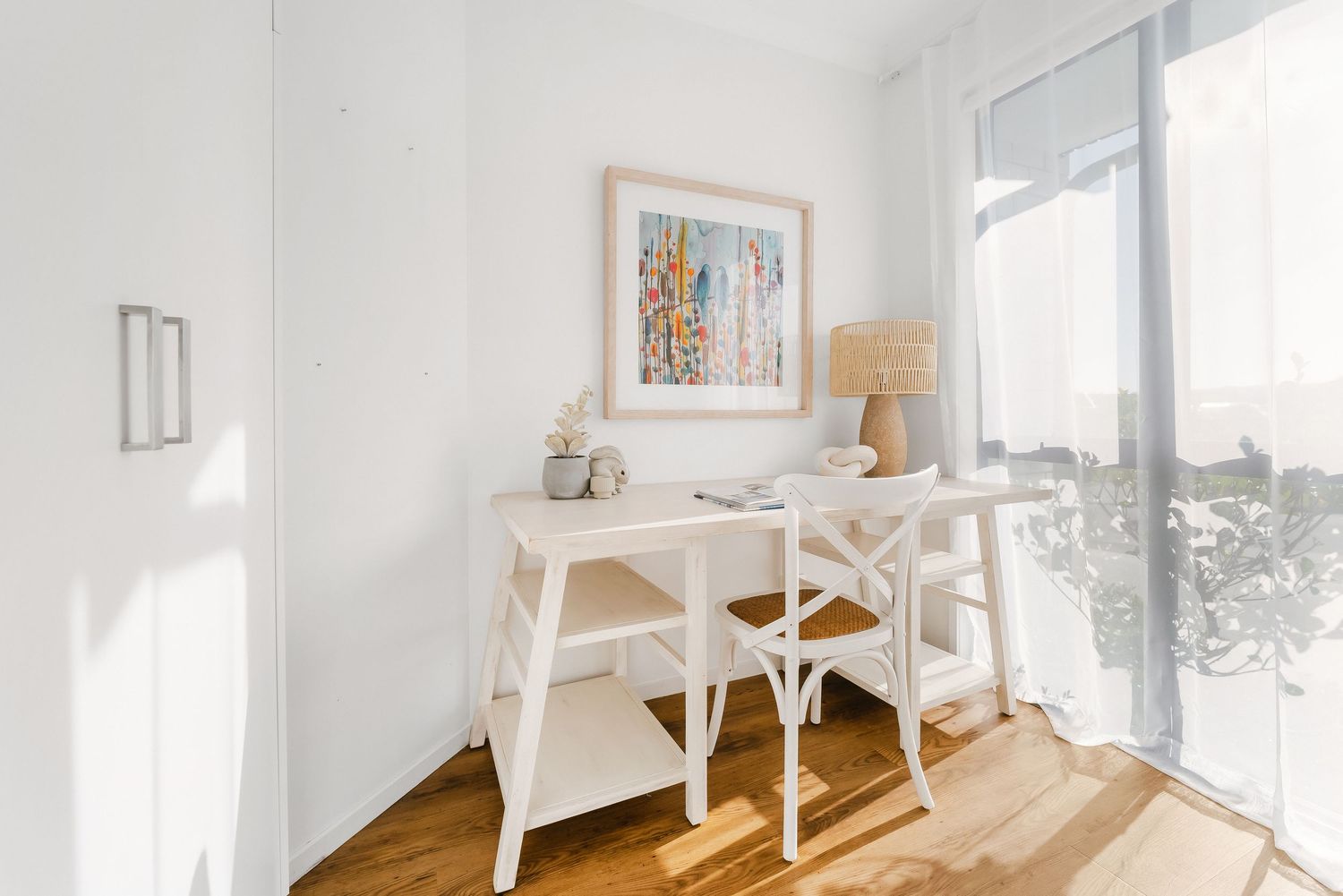 White minimalist home office space with wooden desk, chairs and abstract wall art bathed in natural sunlight.