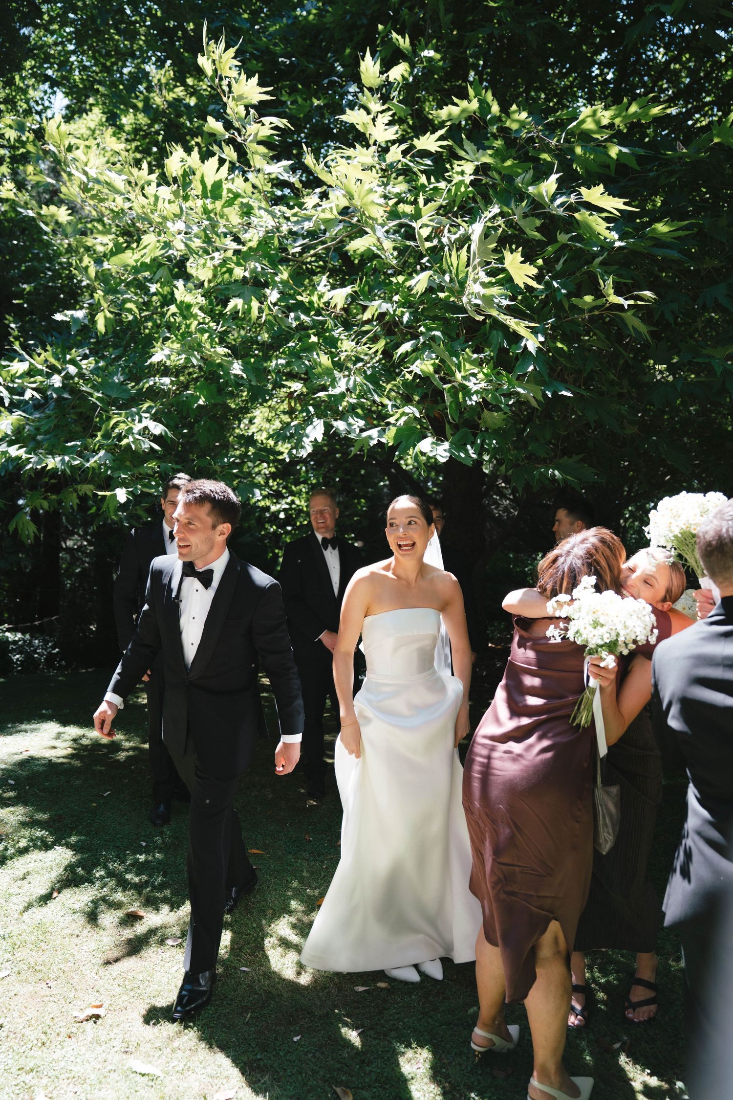 A couple walks along a garden path during an outdoor wedding celebration with guests trailing behind in dappled sunlight.