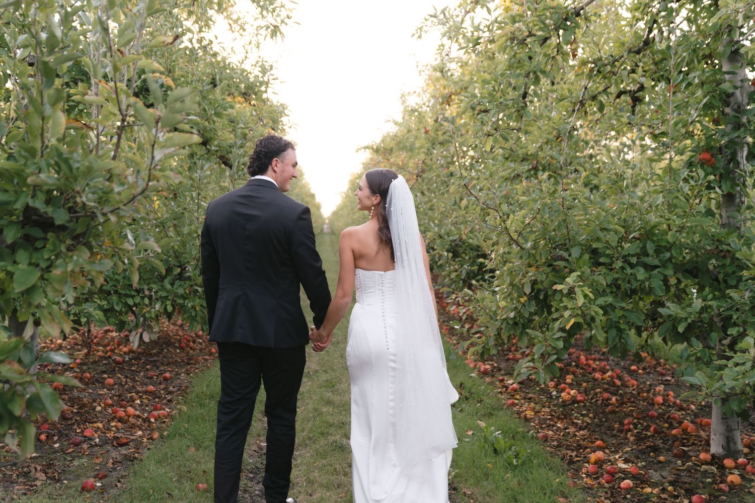 Couple walking through an orchard with fallen autumn leaves on the ground.