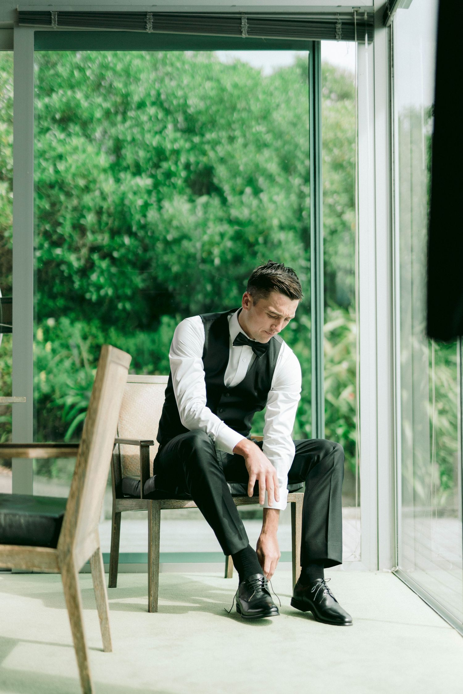 Person in formal vest and dress shirt sitting thoughtfully by large window with green view.