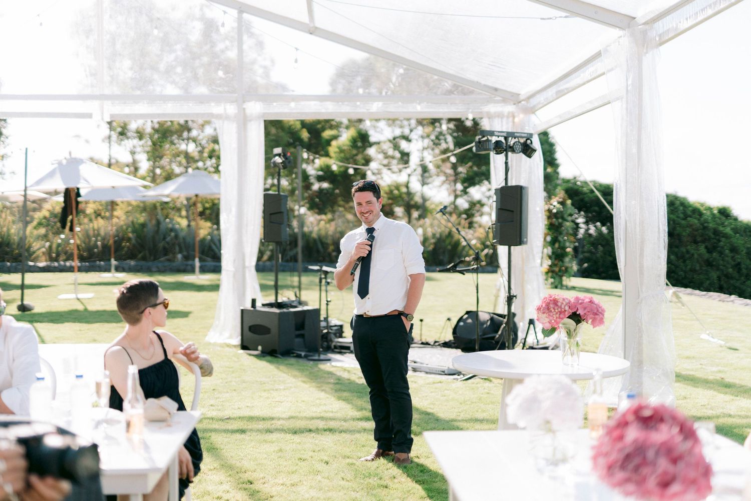 Wedding speaker addresses guests under a white outdoor pavilion surrounded by greenery.