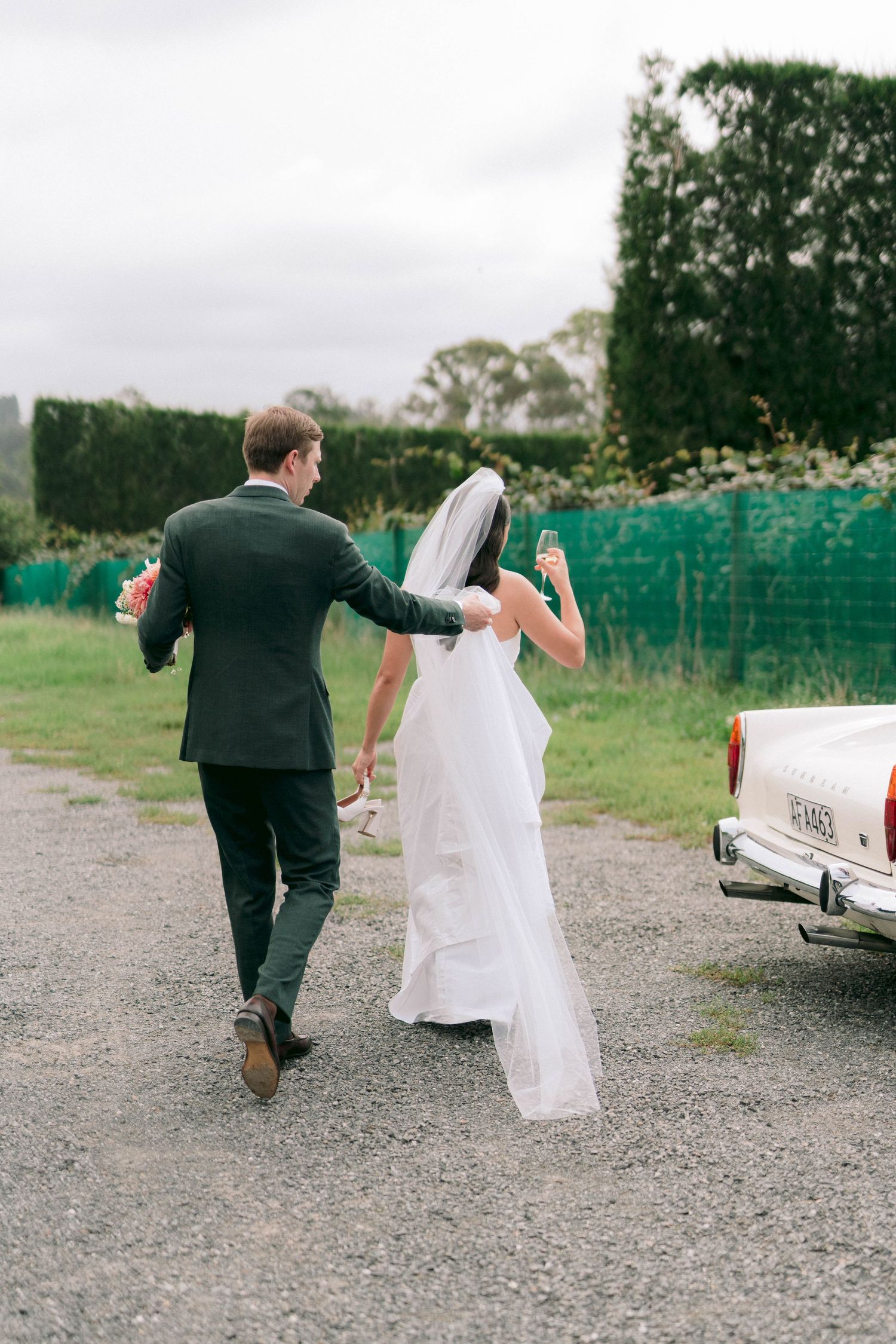 Wedding couple playfully walking on a gravel path near parked cars.