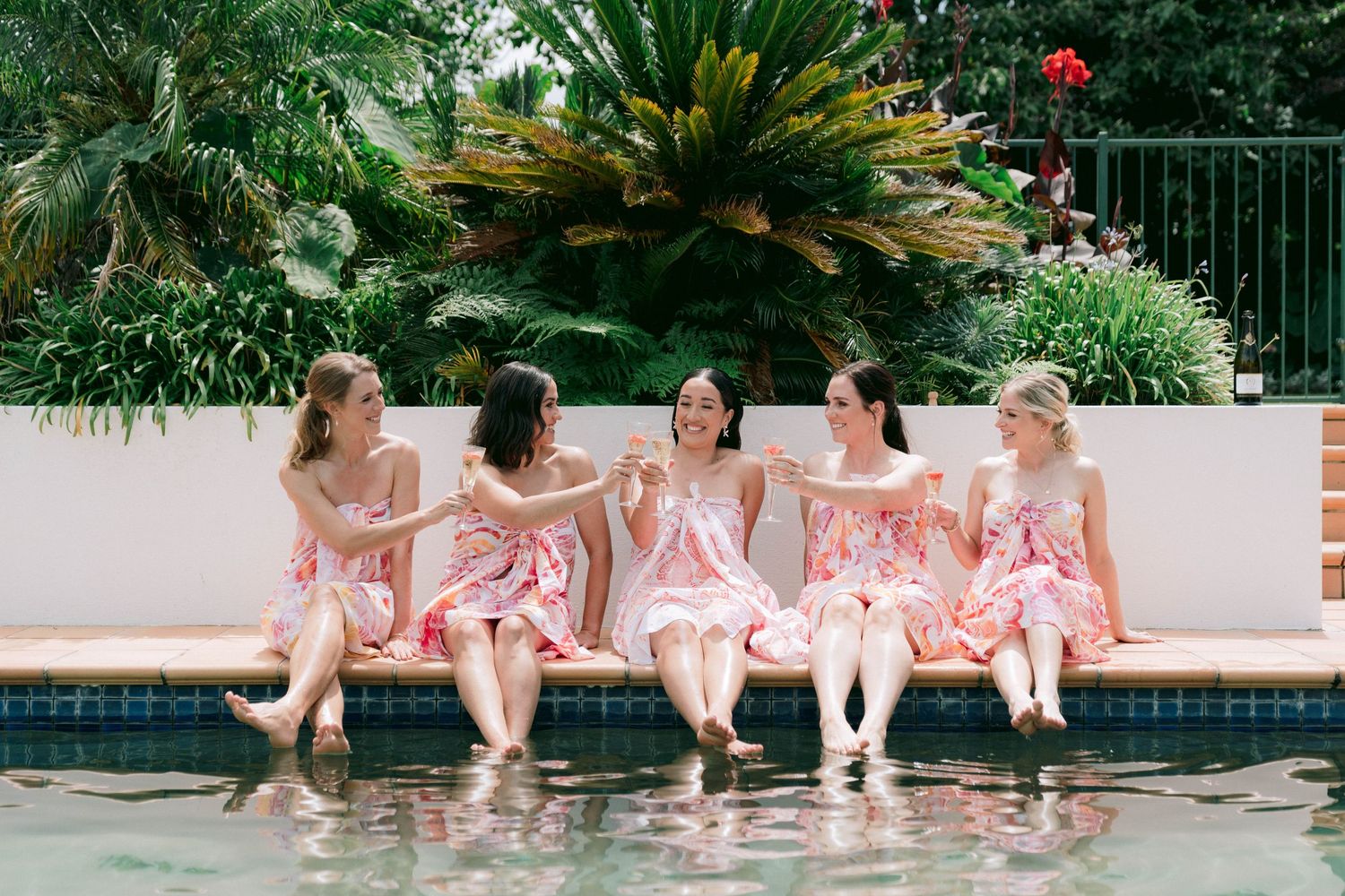 Five people in matching pink outfits sit at the edge of a pool with their feet in the water.