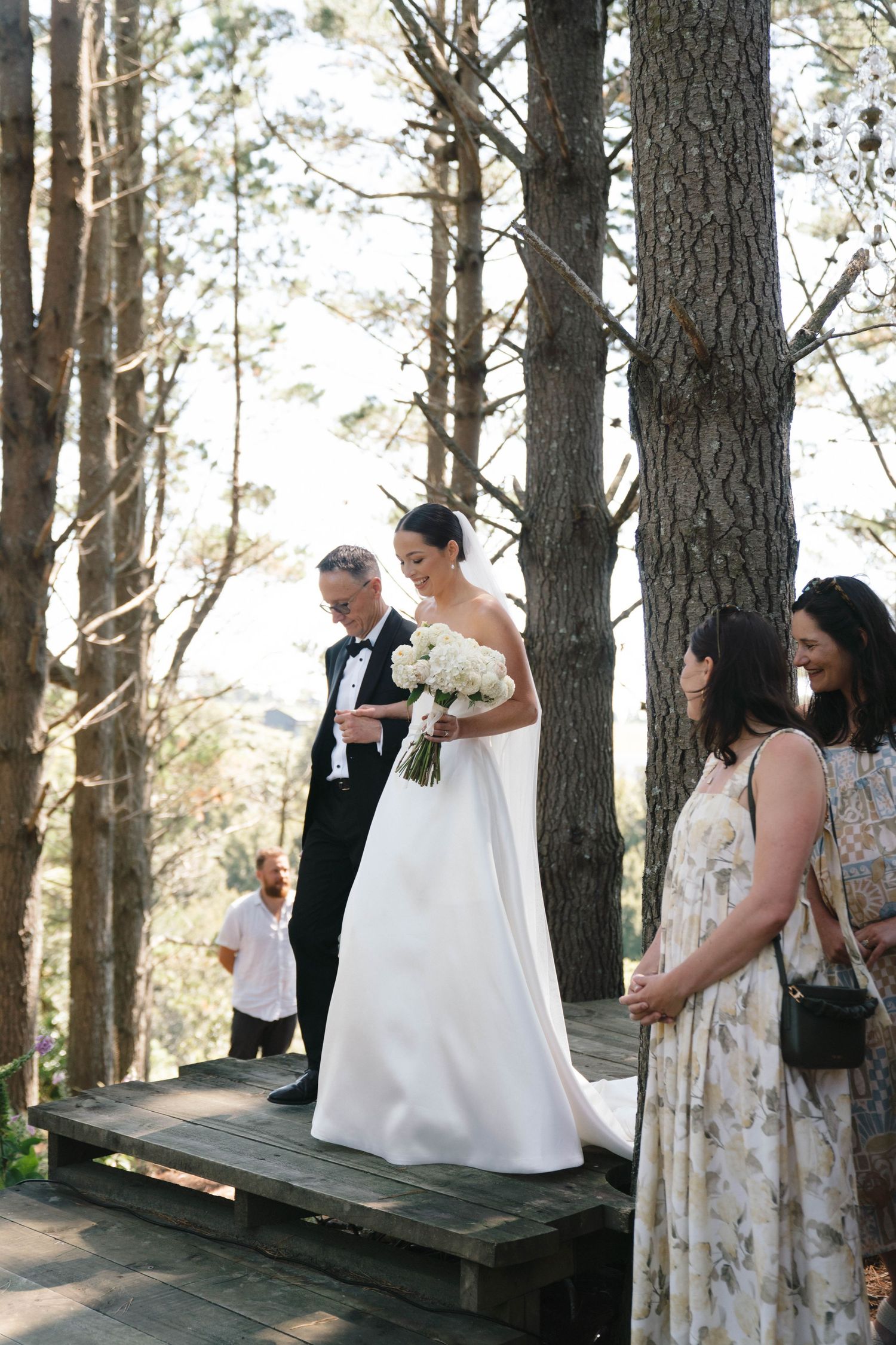 A bride in elegant white gown stands on wooden platform in a forest setting during ceremony.