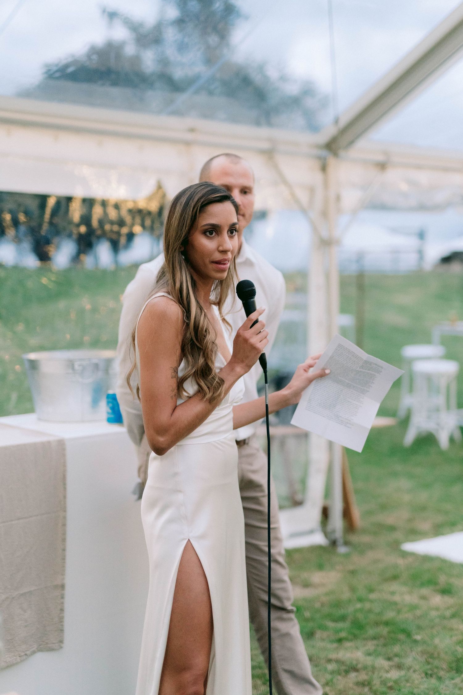 Someone in a sleek white dress with a thigh-high slit delivers a speech at an outdoor wedding reception under a tent.