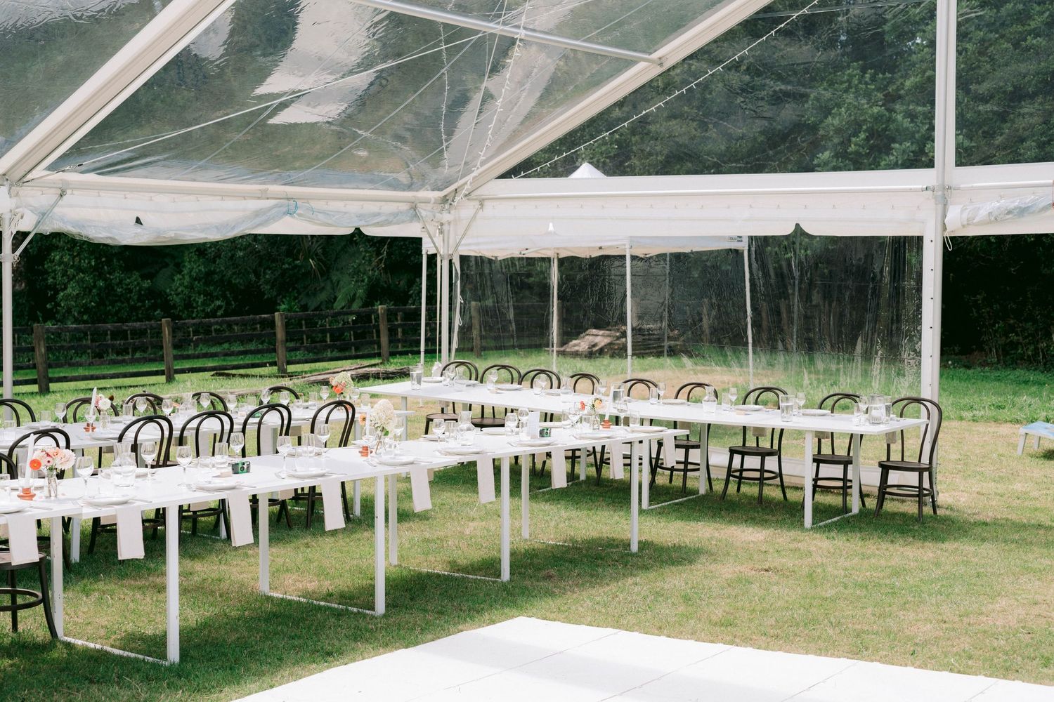 Tables and chairs set up under a clear outdoor tent canopy for a wedding reception on grass with trees in background.