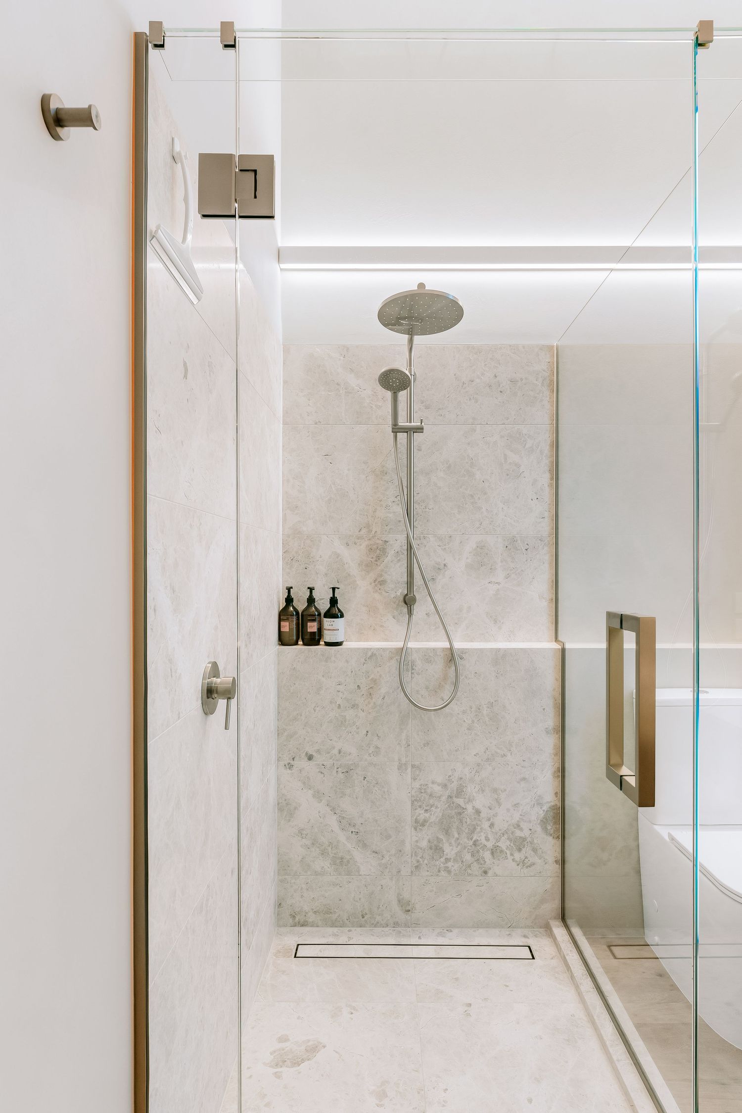 Modern minimalist shower enclosure with glass doors, marble-look tiles, and brushed gold fixtures featuring a rainfall showerhead.