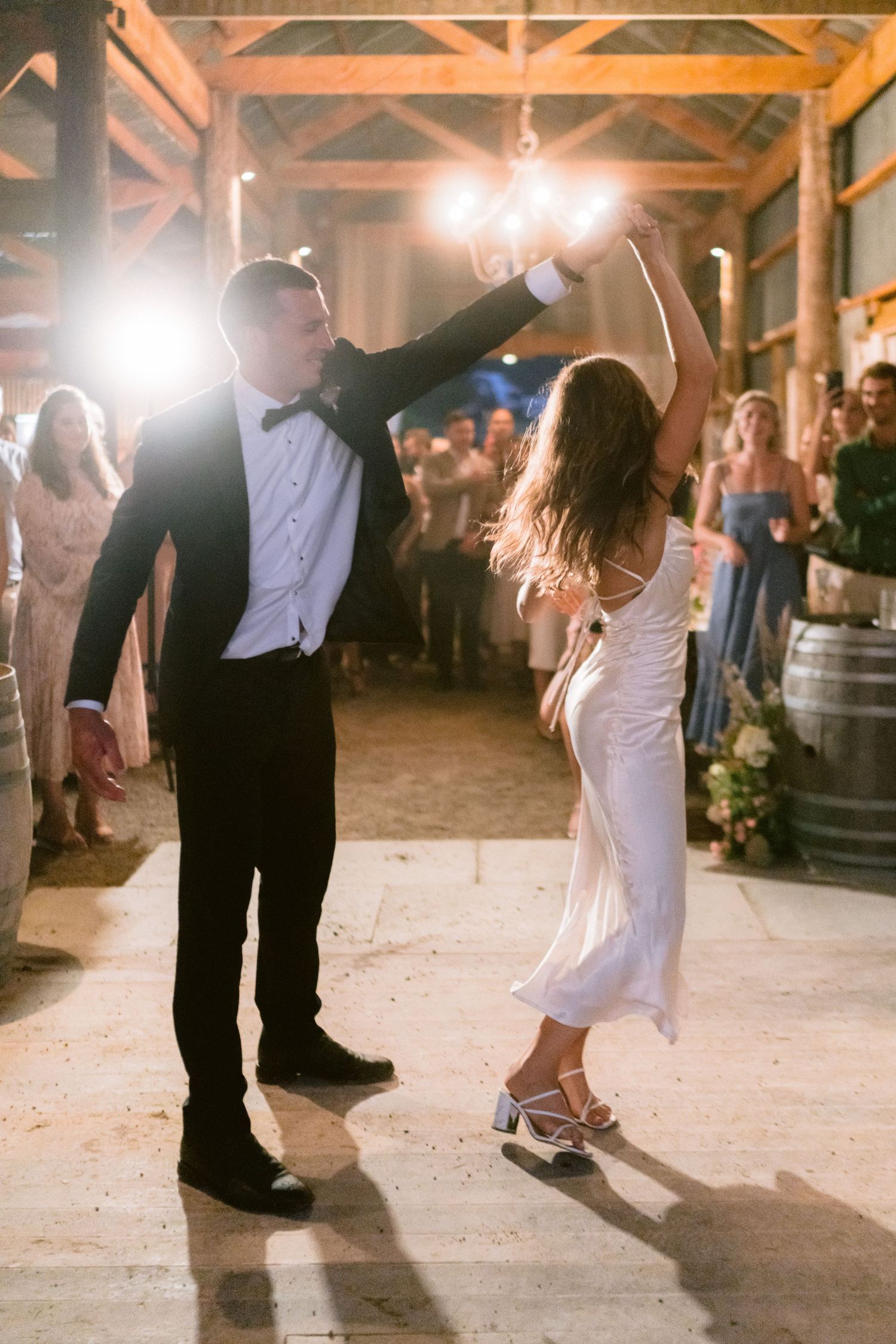 Couples dancing on rustic barn floor during wedding reception with dramatic lighting and barrel decor.