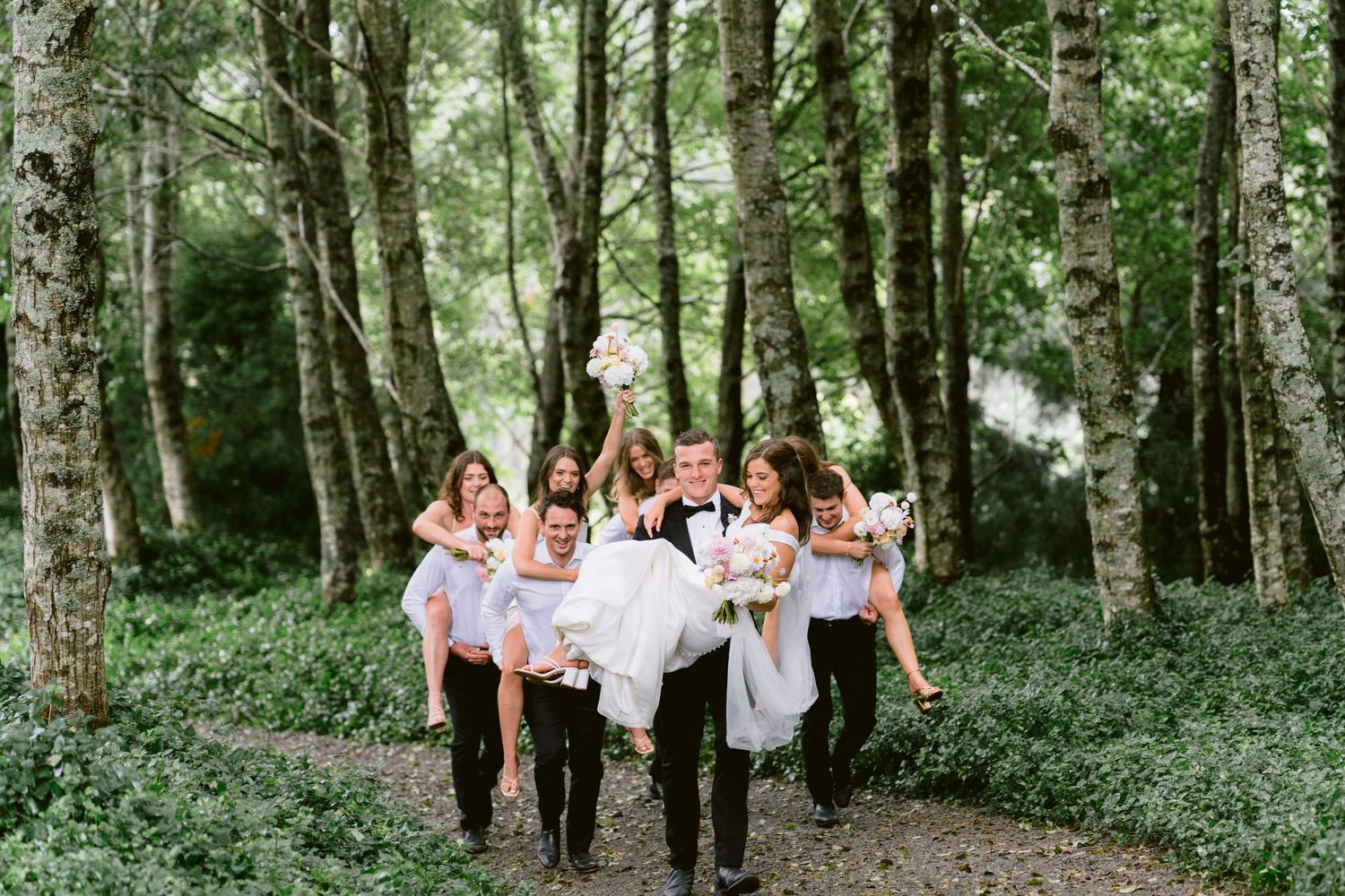 Wedding party carries bride through a forest path while laughing and celebrating.