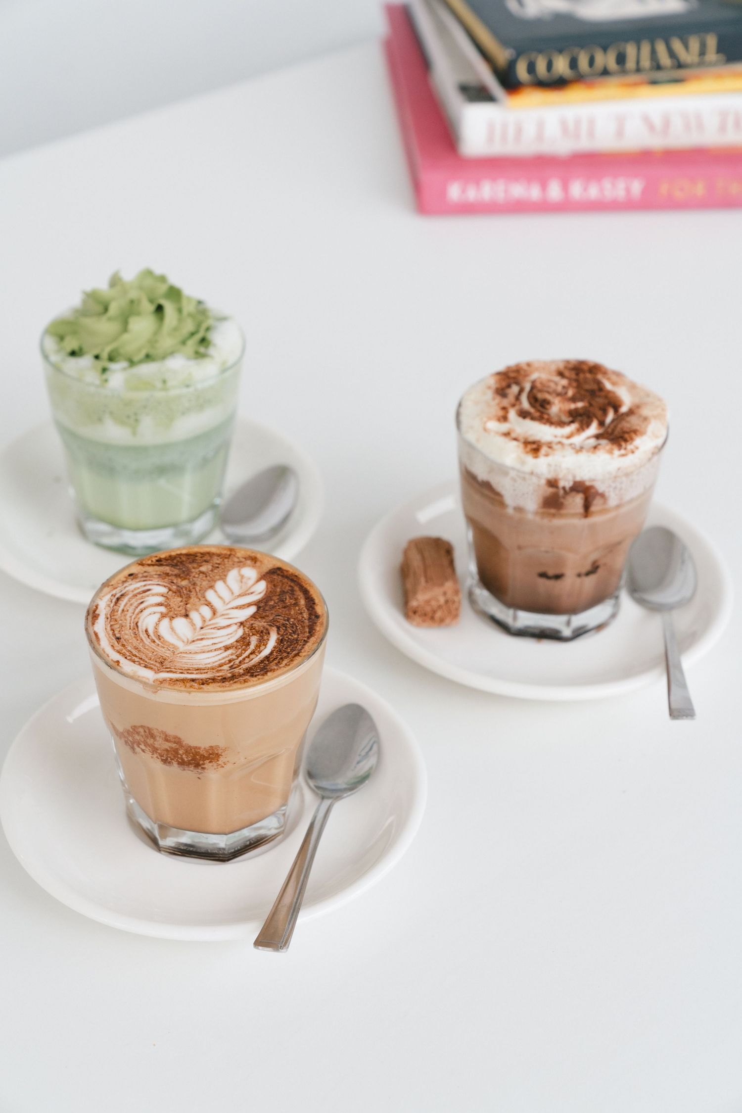 Three artisanal coffee drinks in clear glasses with latte art, including a matcha latte and two chocolate beverages on white.