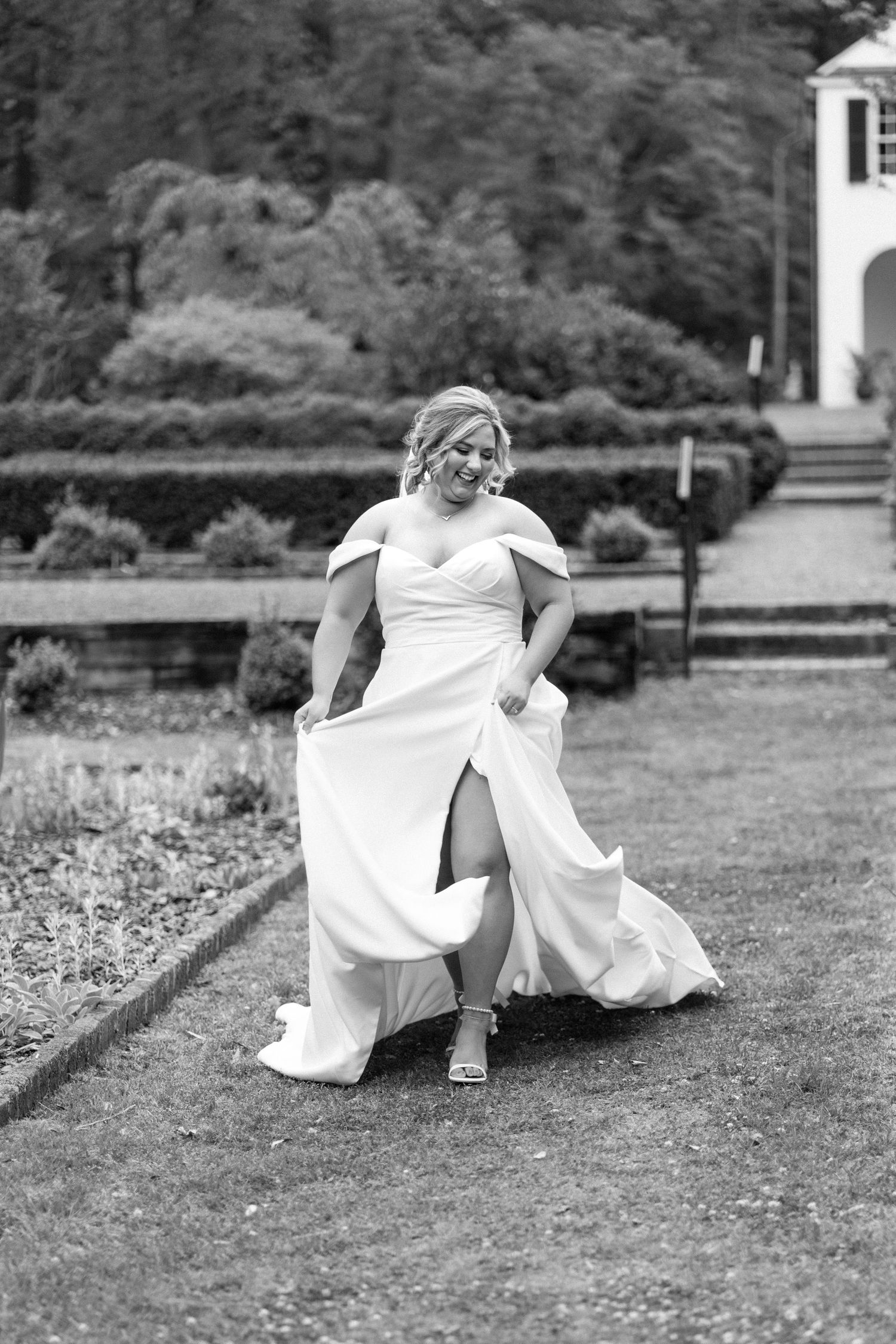 A bride in a flowing white dress dances joyfully on a garden lawn with a church building visible in the background.