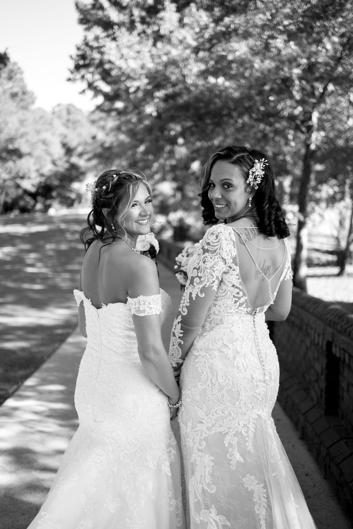 Two brides in elegant lace gowns pose together outdoors in black and white.