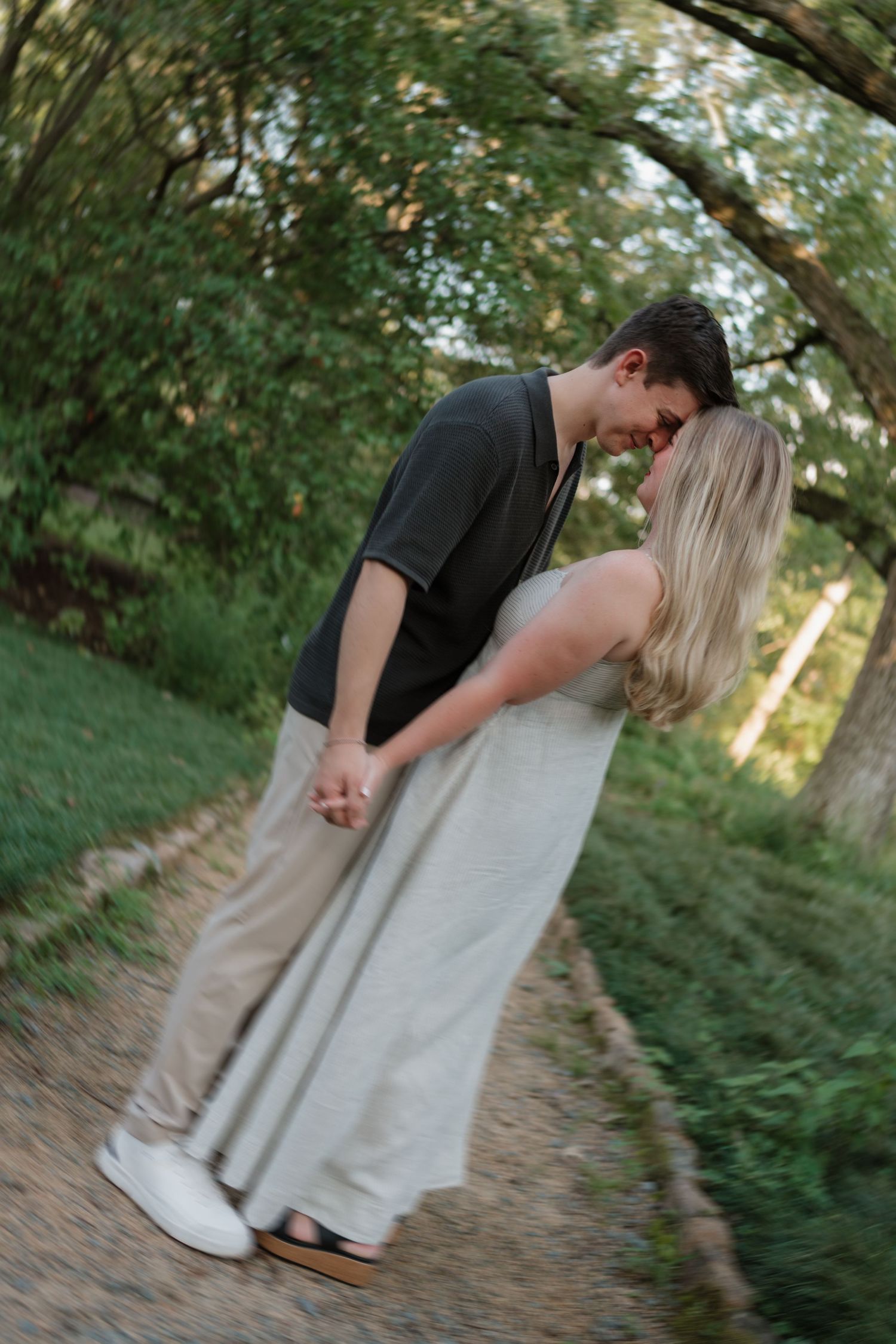 A couple shares a romantic moment on a tree-lined path, with the person in gray dress leaning back while holding hands.