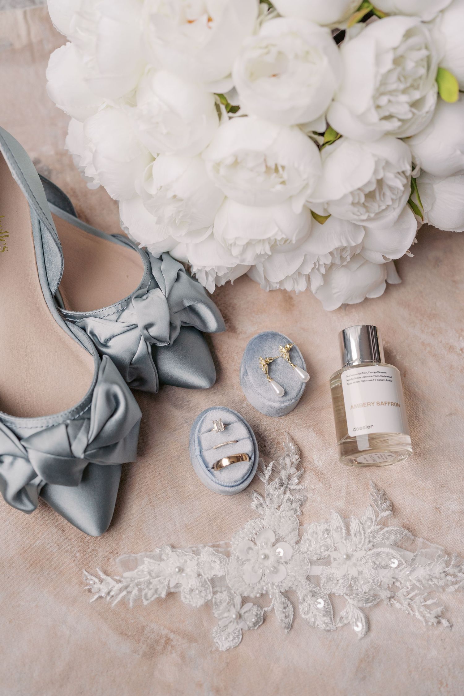 Wedding accessories including blue bow shoes, rings, perfume and white peonies arranged on beige surface.