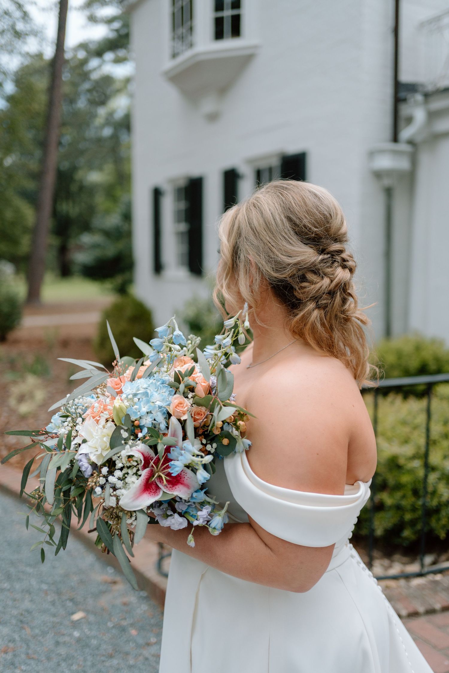 Elegant bridal bouquet with blue and peach flowers held by bride in off-shoulder white dress against white building exterior.