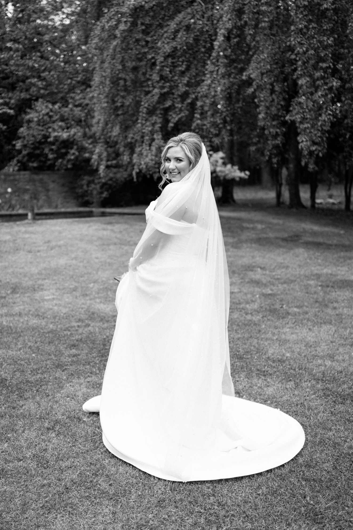 Black and white portrait of a bride in flowing gown standing on grass beneath weeping willow trees.