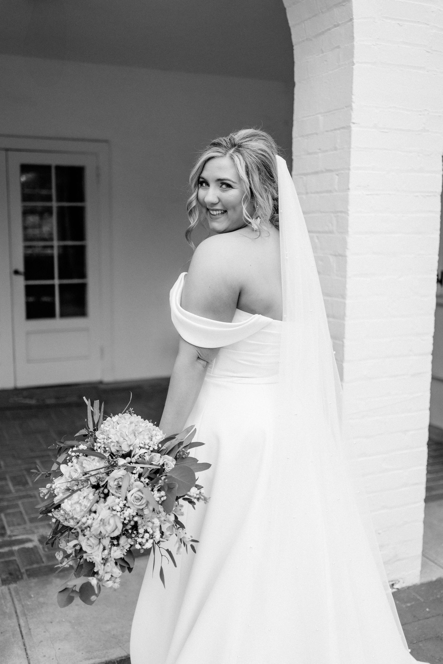 Black and white bridal portrait showing off-shoulder gown details and flowing veil under archway.