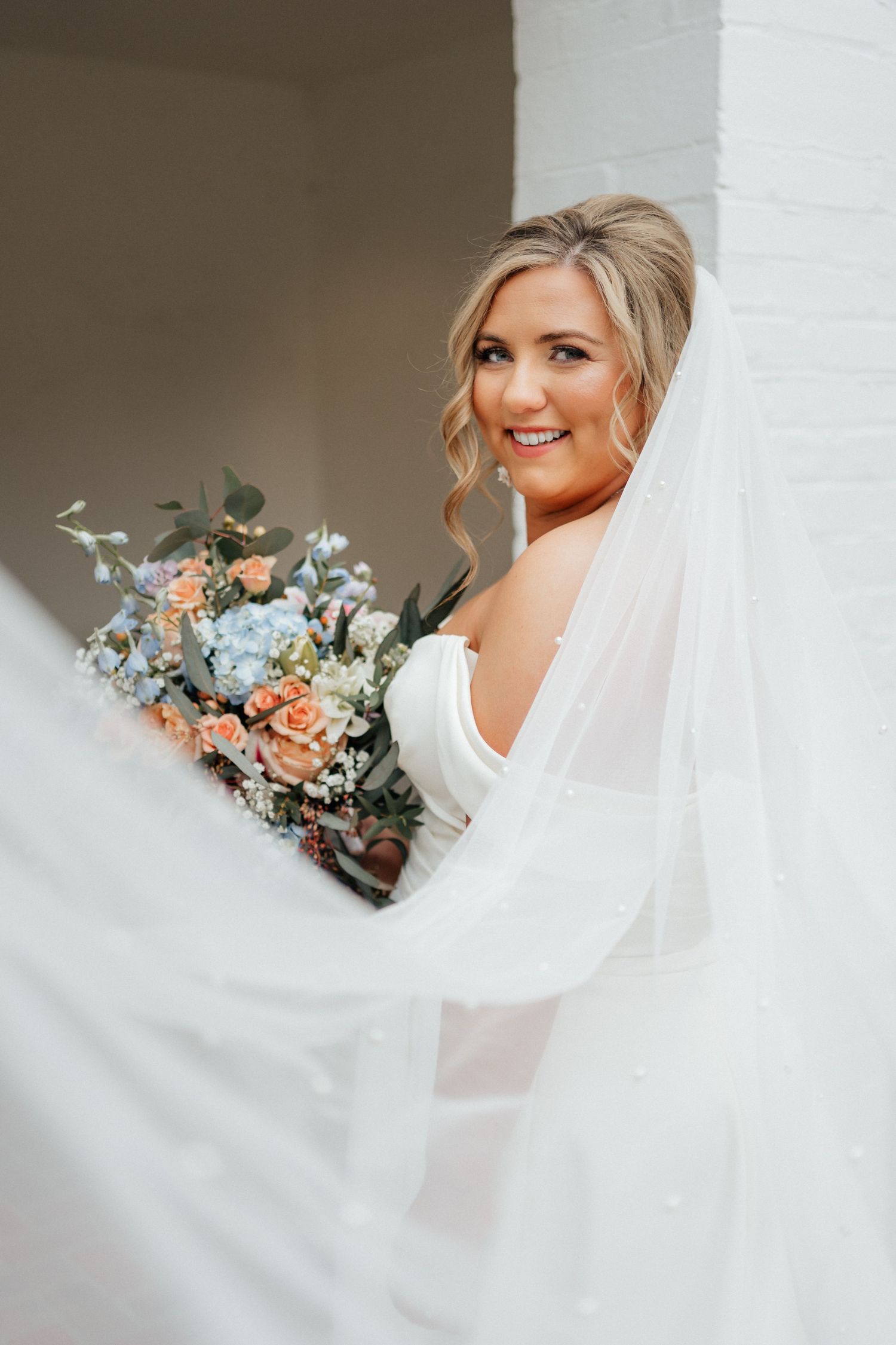 Elegant bridal portrait featuring a flowing veil and romantic peach and blue floral bouquet against white architecture.