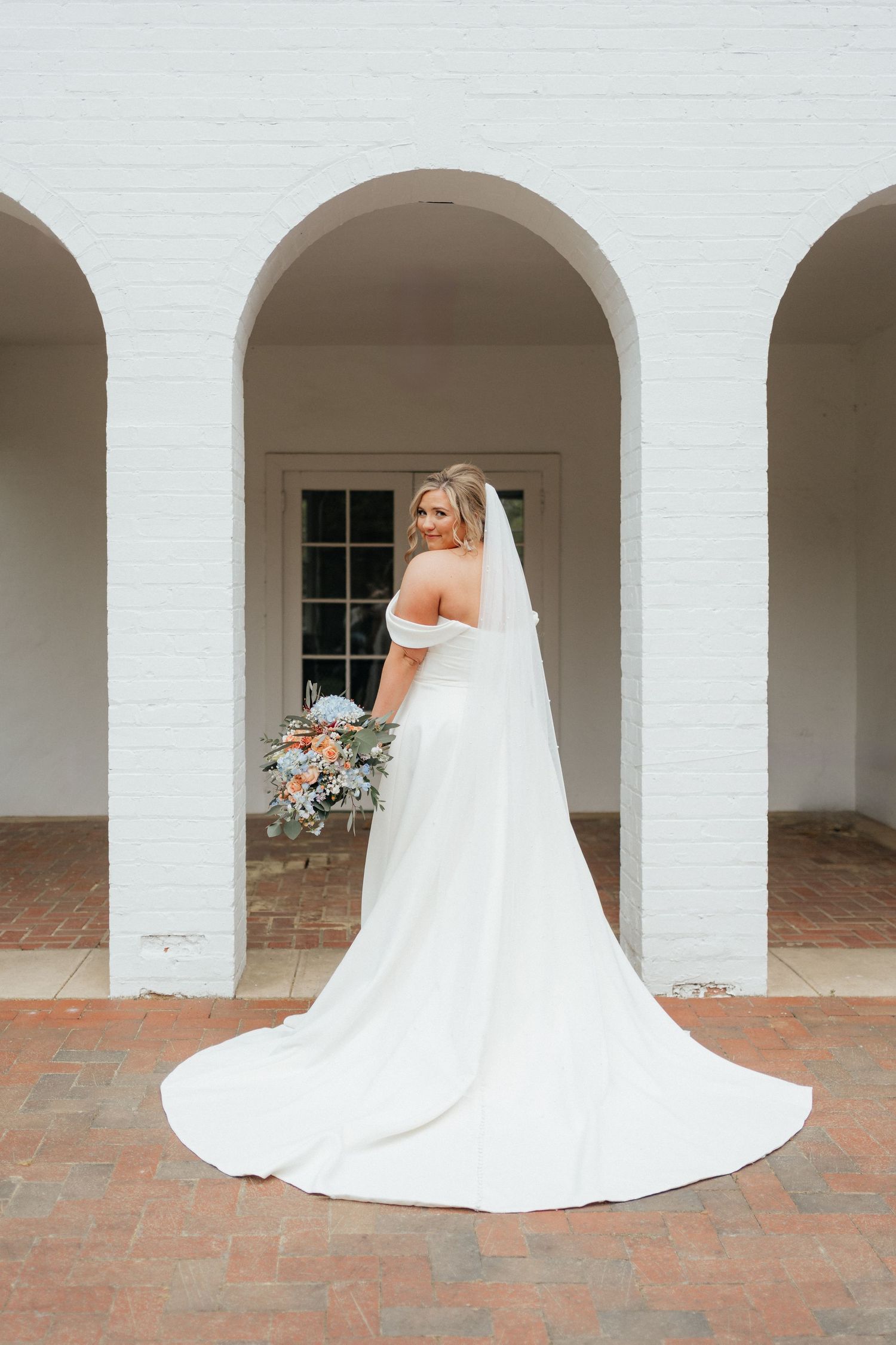 Elegant bride in flowing white gown poses between classic arches holding a bouquet against white walls.