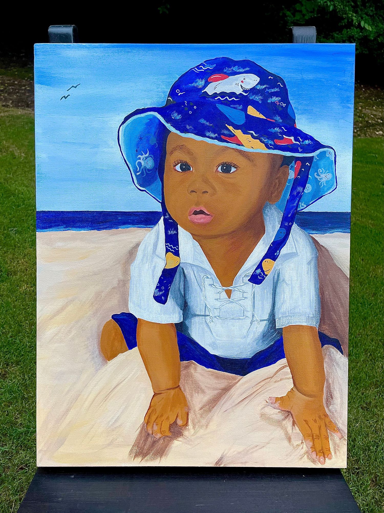 Hand painted Acrylic on canvas painting of a young son in blue hat sitting on beach with ocean background.