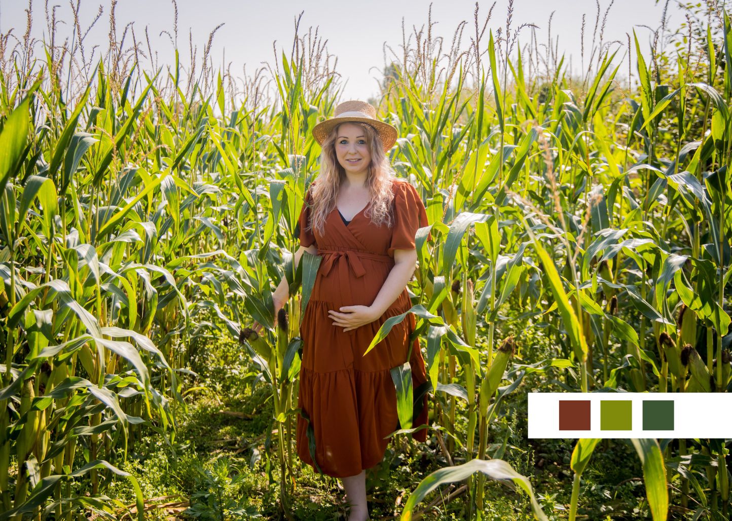Woman in rust-colored dress standing in tall green cornfield wearing straw hat.
