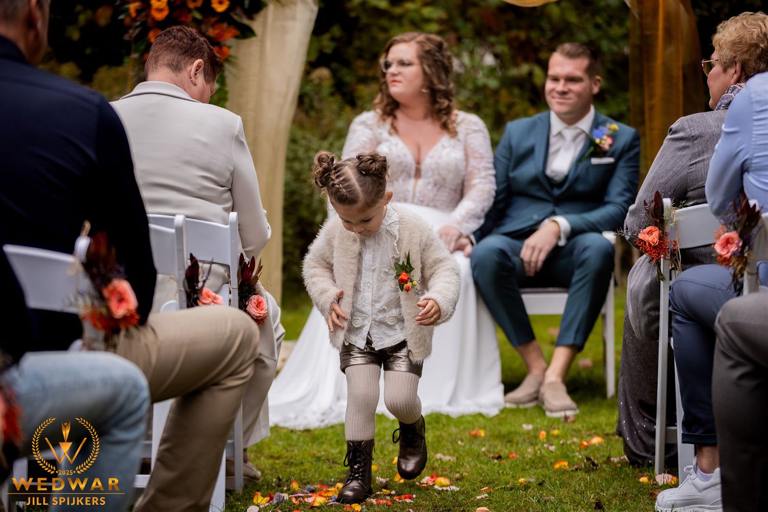 A young flower girl in a white dress walks down the outdoor wedding aisle.