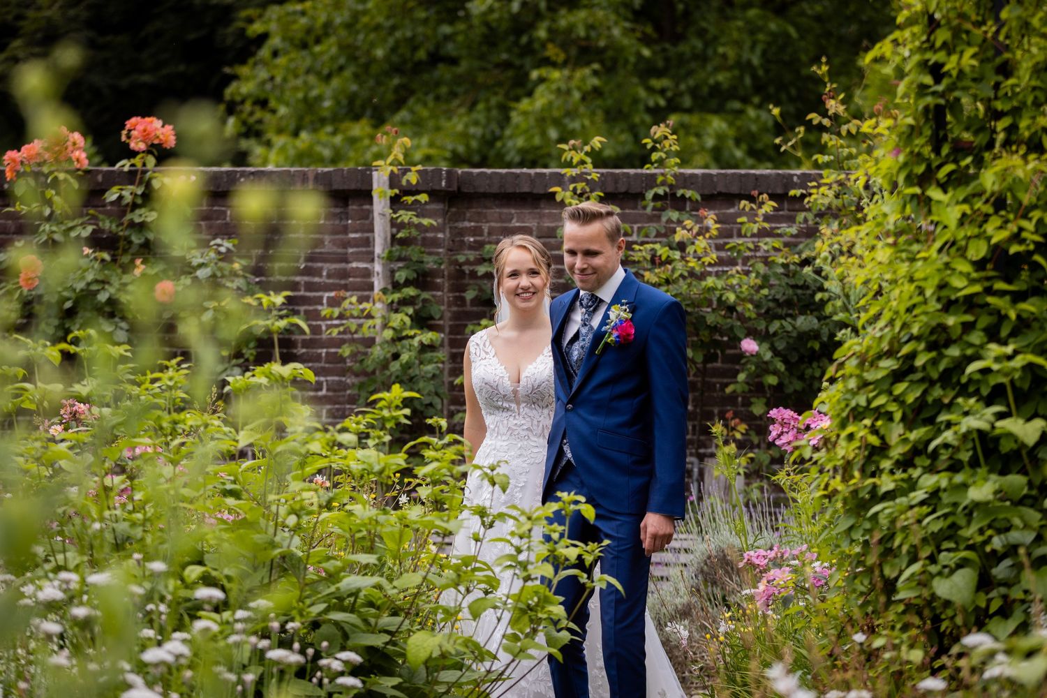 Couple posing in beautiful garden setting with pink flowers and stone wall on their wedding day.