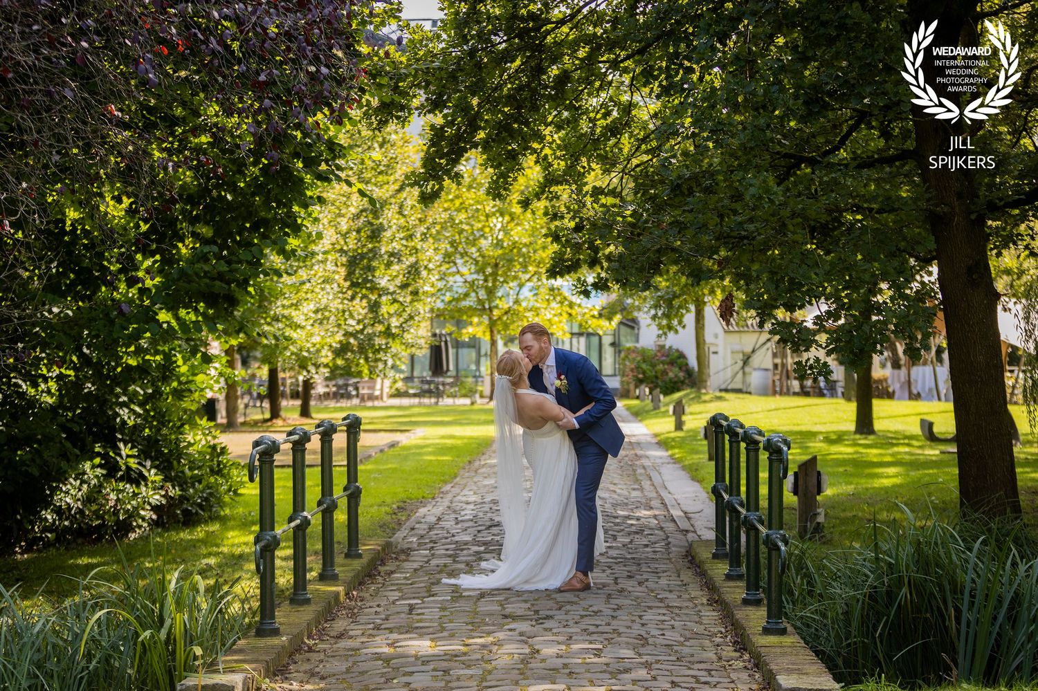 Newlyweds pose on a garden pathway lined with trees and iron fencing.