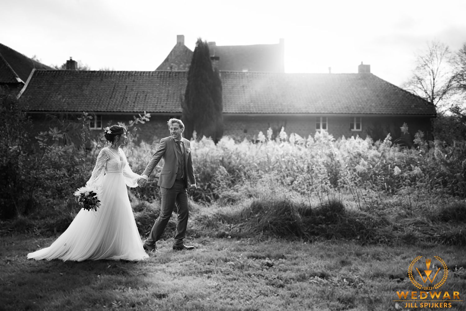 Black and white wedding photo of couple walking through sunlit garden path.