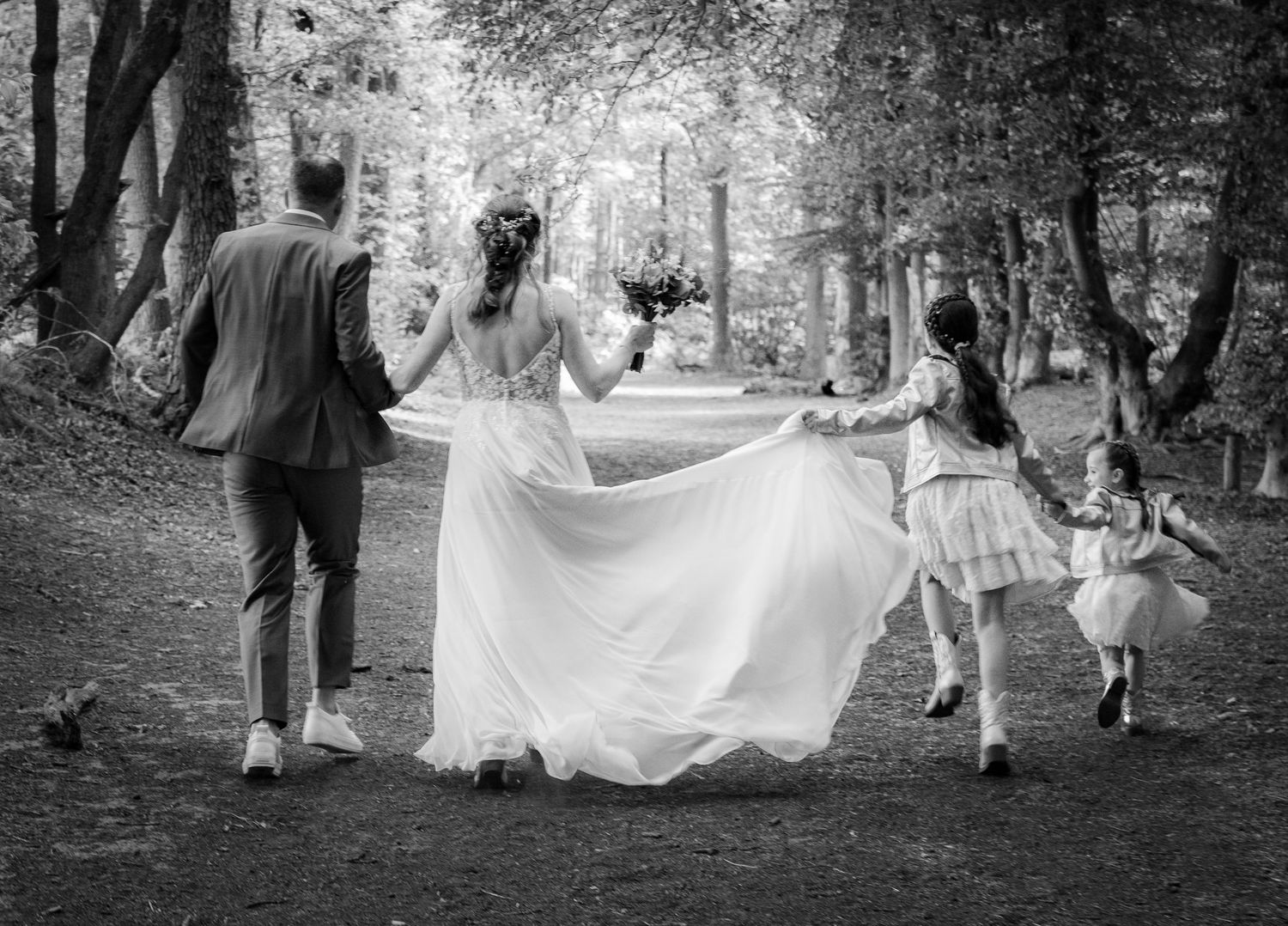 Black and white photo of family walking through tree-lined path with flowing wedding dress.