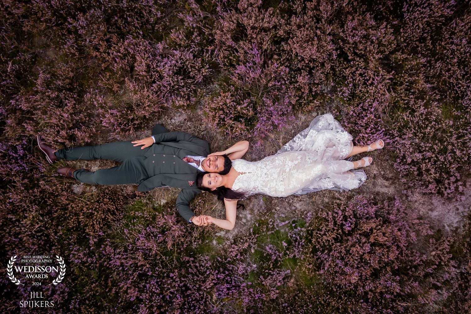 Aerial view of couple lying in field of purple flowers wearing wedding attire.